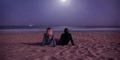 a man and a woman are sitting on the beach looking at the moon .