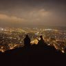 two people are sitting on top of a hill overlooking a city at night .