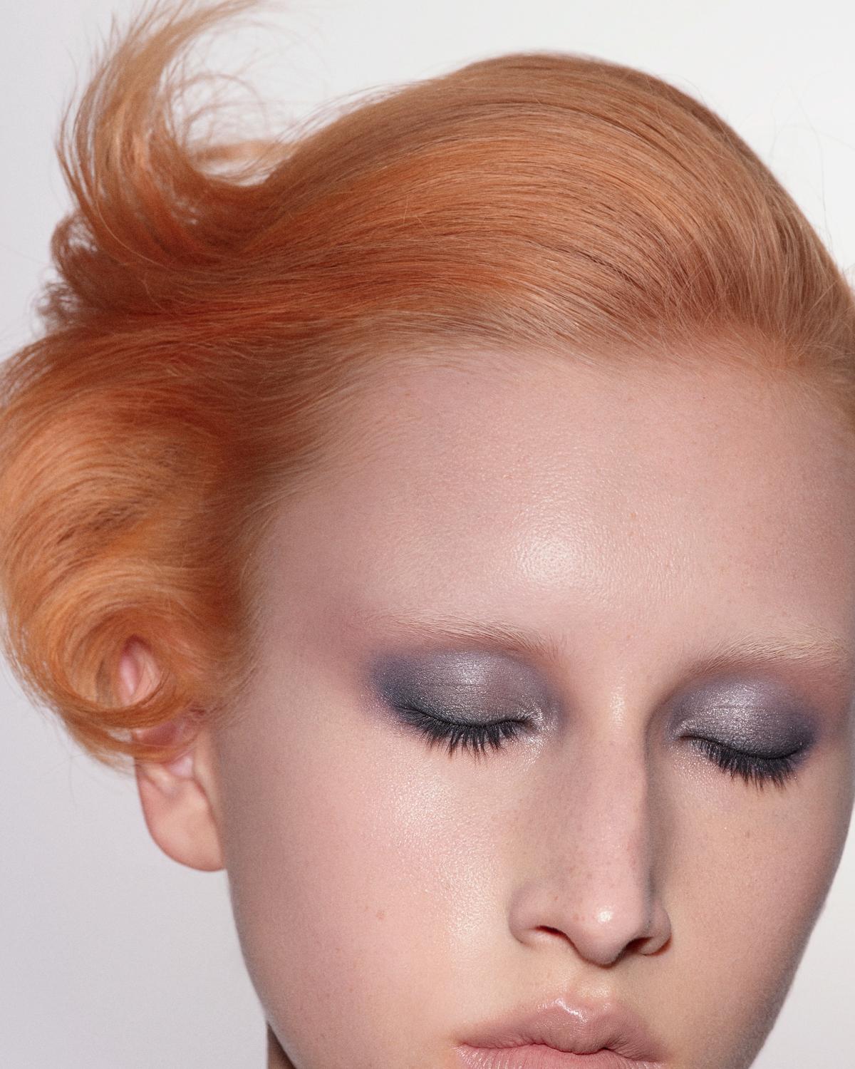 Close-up of a woman with closed eyes, smoky gray-violet eyeshadow, and swept-up copper-orange hair against a white background.