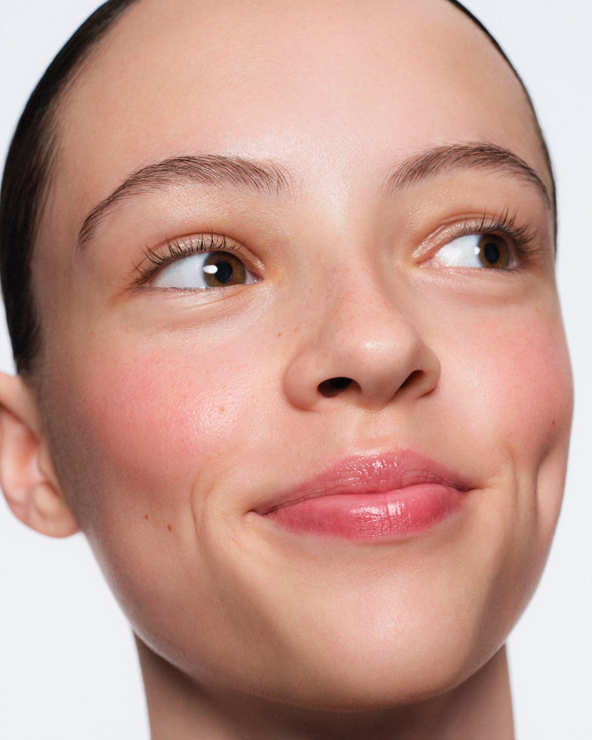 Close-up of a woman with slicked-back hair, glancing sideways with a soft smile, rosy blush, light freckles, and glossy pink lips.