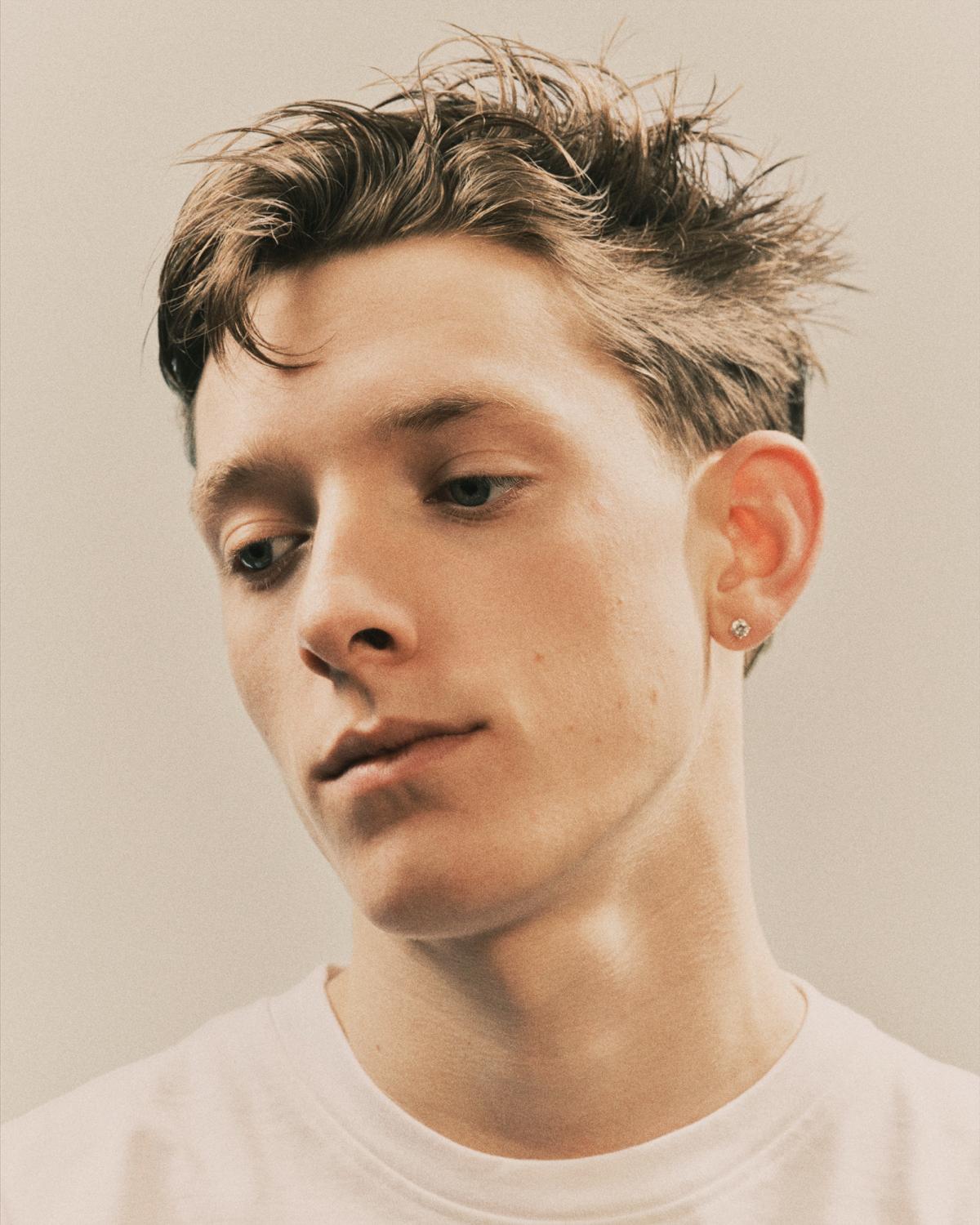 Portrait of a young man with tousled, wet-look brown hair and a diamond stud earring, wearing a white tee and looking down with his head tilted