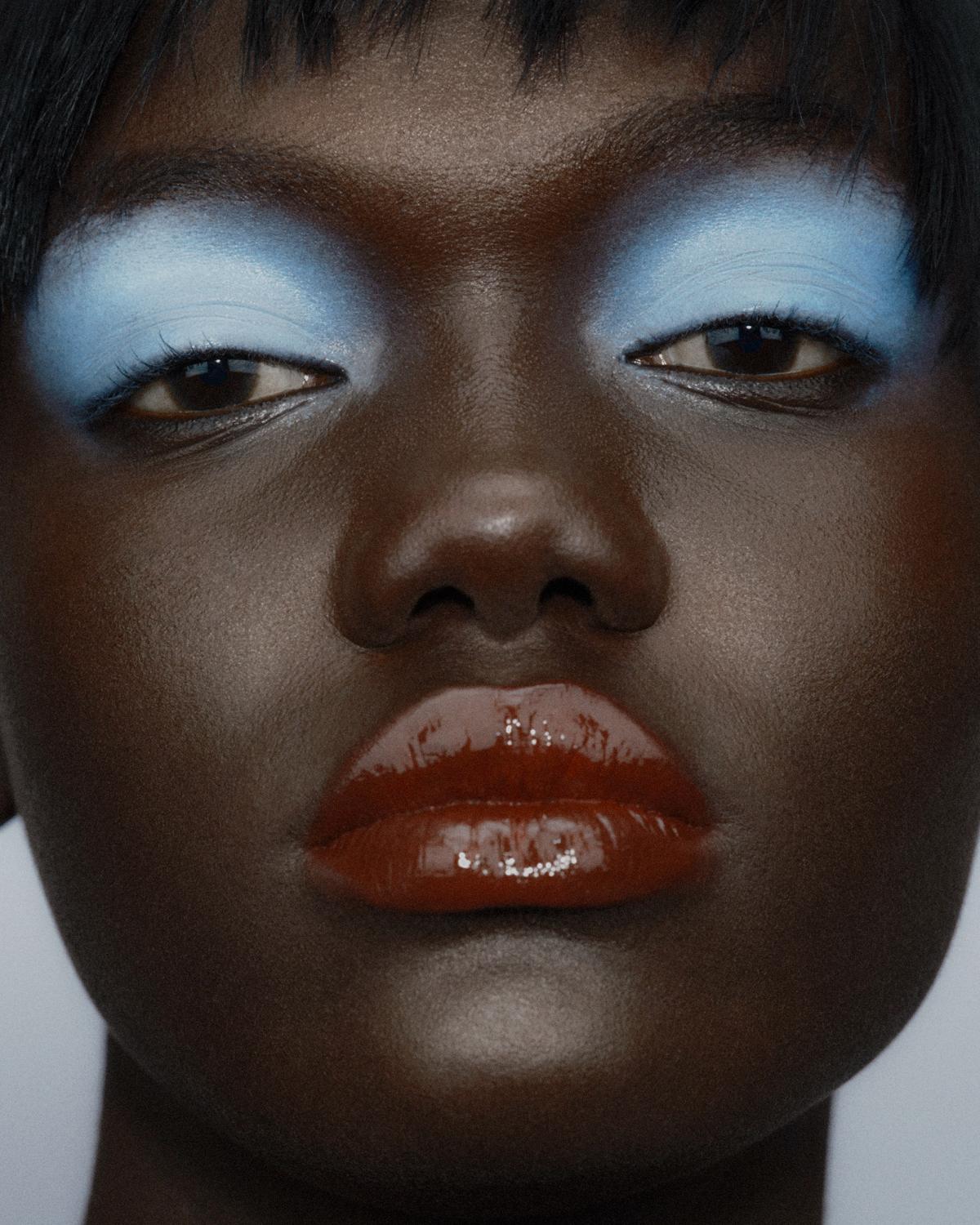 Close-up of a woman with bold powder-blue eyeshadow and high-shine rust-red lips, looking at the camera with glowing skin and short black bangs.