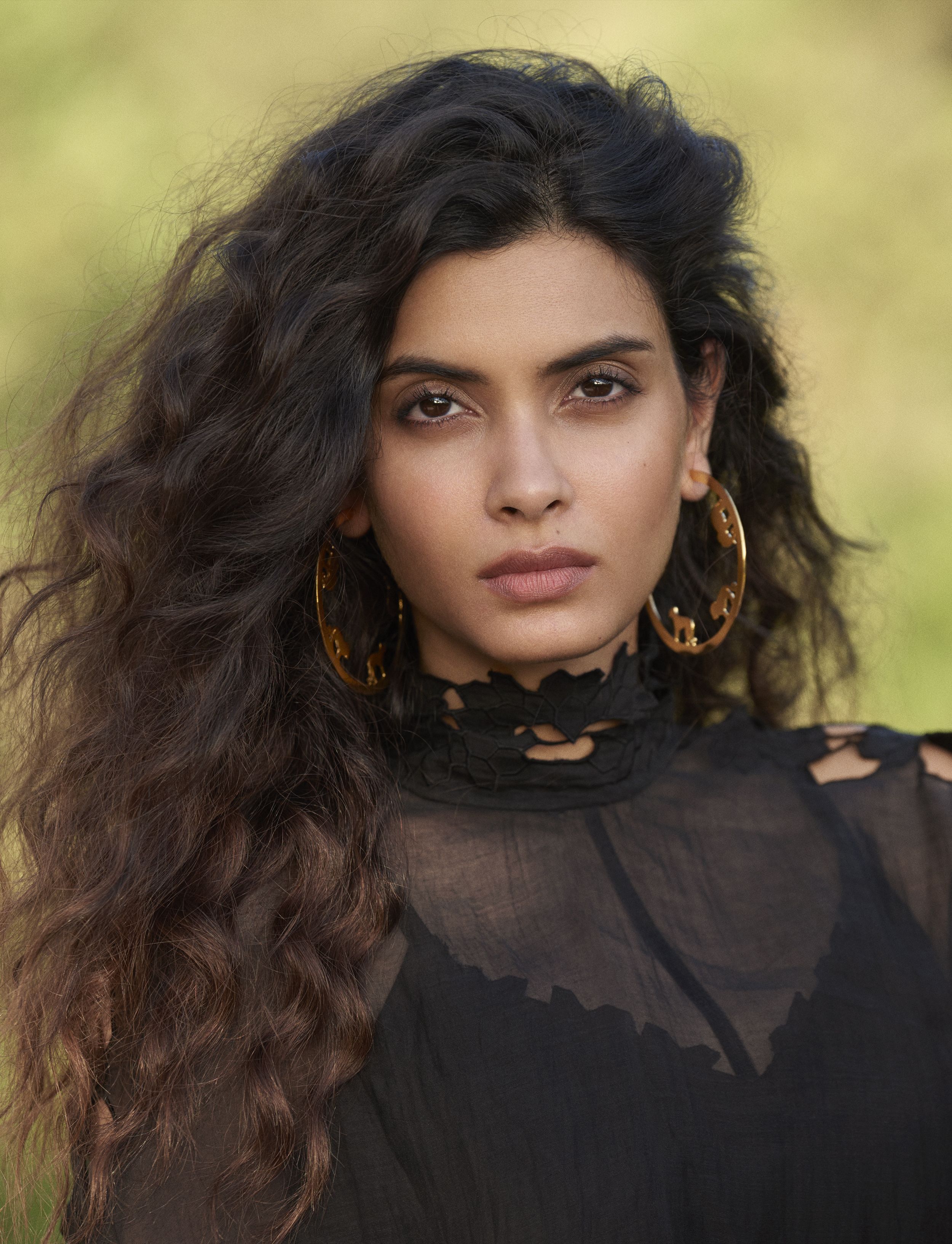 Diana Penty with long, voluminous dark waves, large gold animal-motif hoop earrings, and a sheer black lace-collar top, against a soft green background