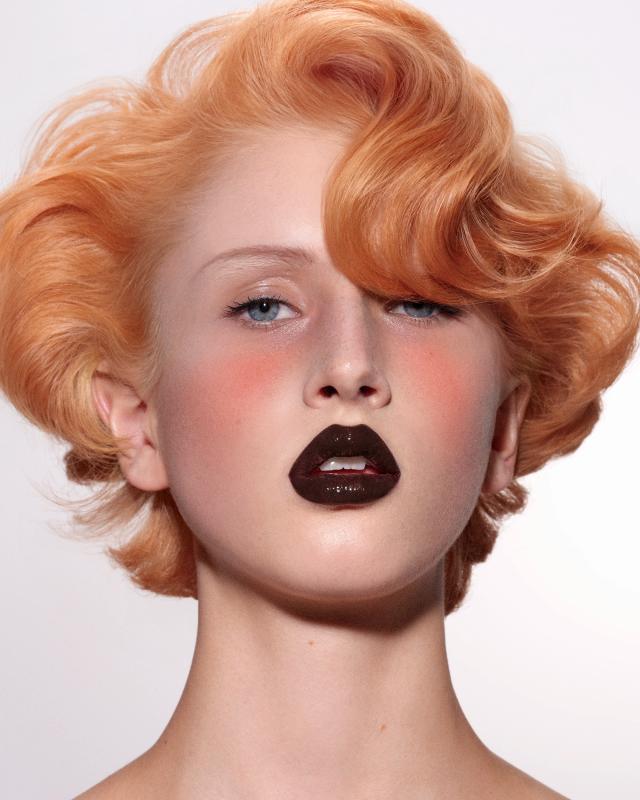 Portrait of a woman with copper-blonde pin curls, rosy blush, and glossy deep chocolate-brown lipstick against a white background