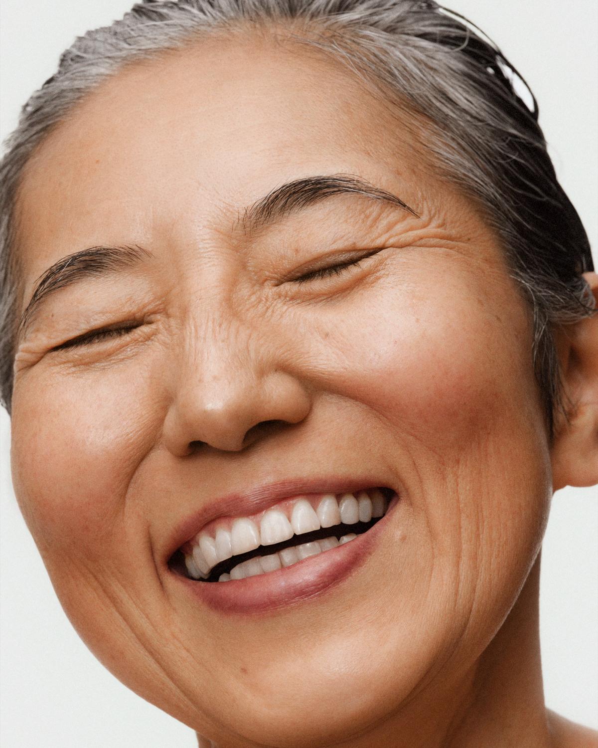 Close-up of an older woman laughing with eyes closed, slicked-back gray hair, natural skin with fine lines, and rosy cheeks