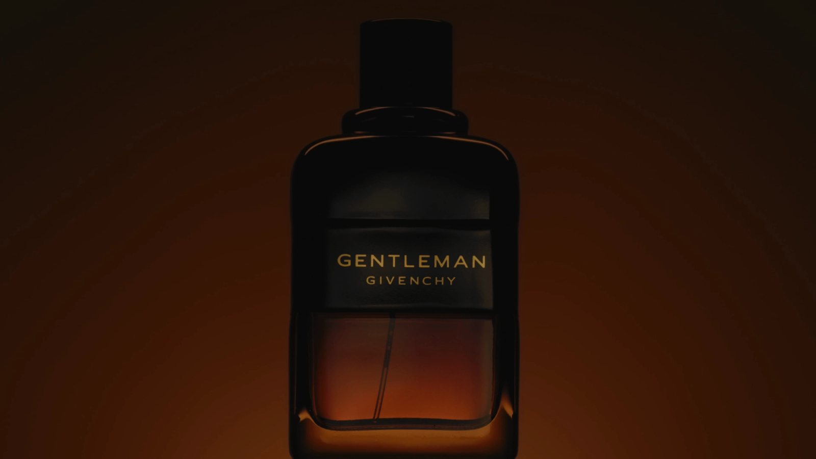 Moody still life of a Givenchy Gentleman perfume bottle with gold lettering, glowing amber against a dark brown background, from a personal video project