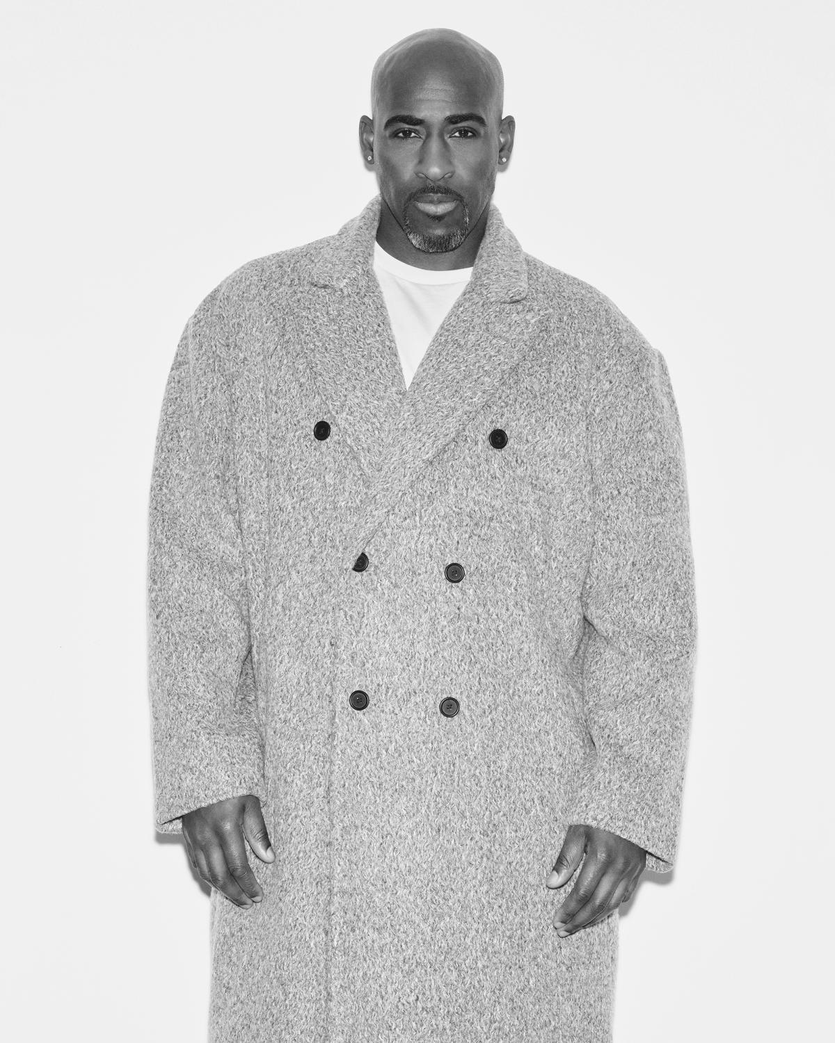 Black-and-white portrait of actor Daniel Francis standing in an oversized double-breasted wool coat over a white tee, with a goatee, facing the camera.