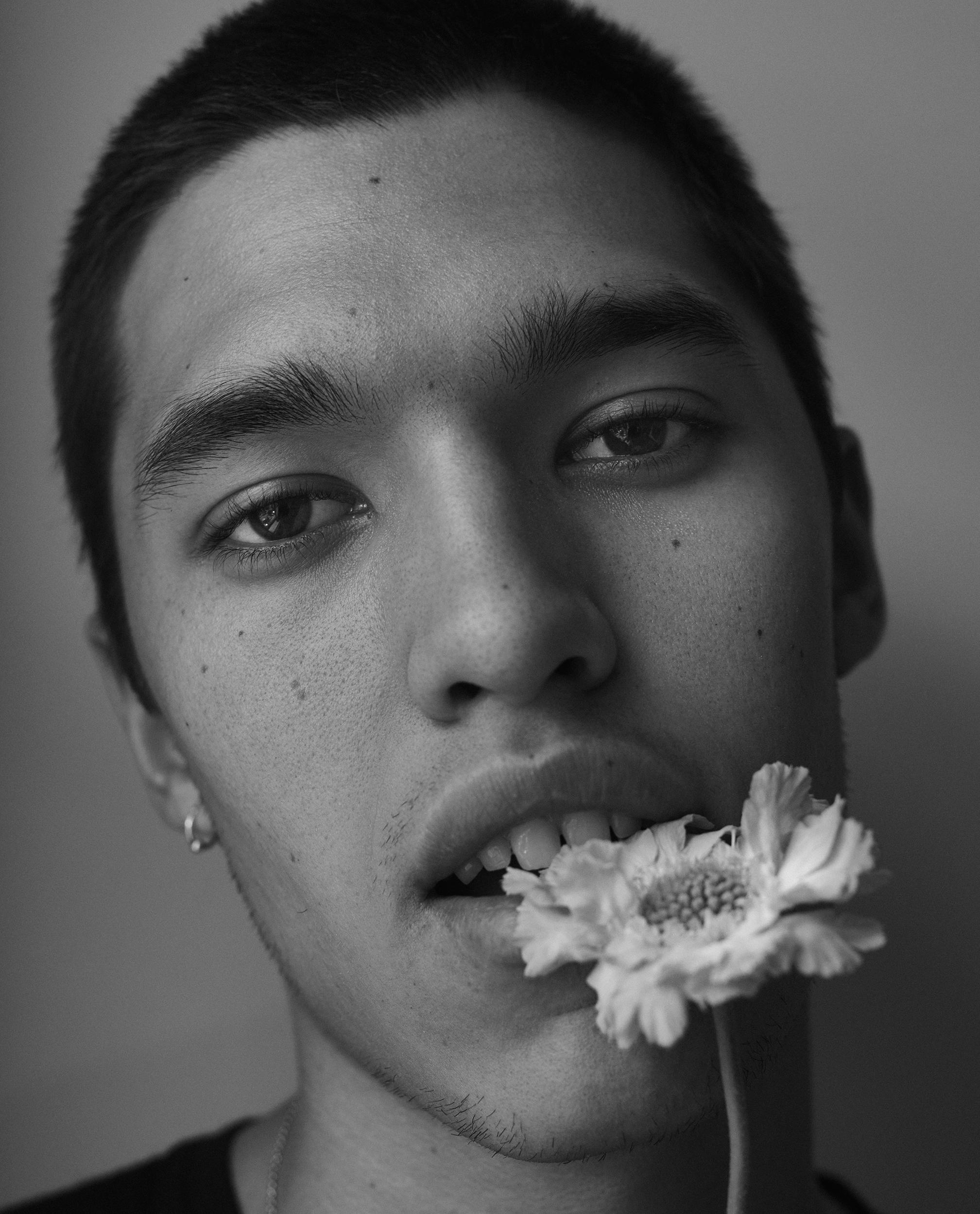 Black-and-white close-up of a young man with a buzz cut and a small hoop earring, looking at the camera with a white flower held between his teeth