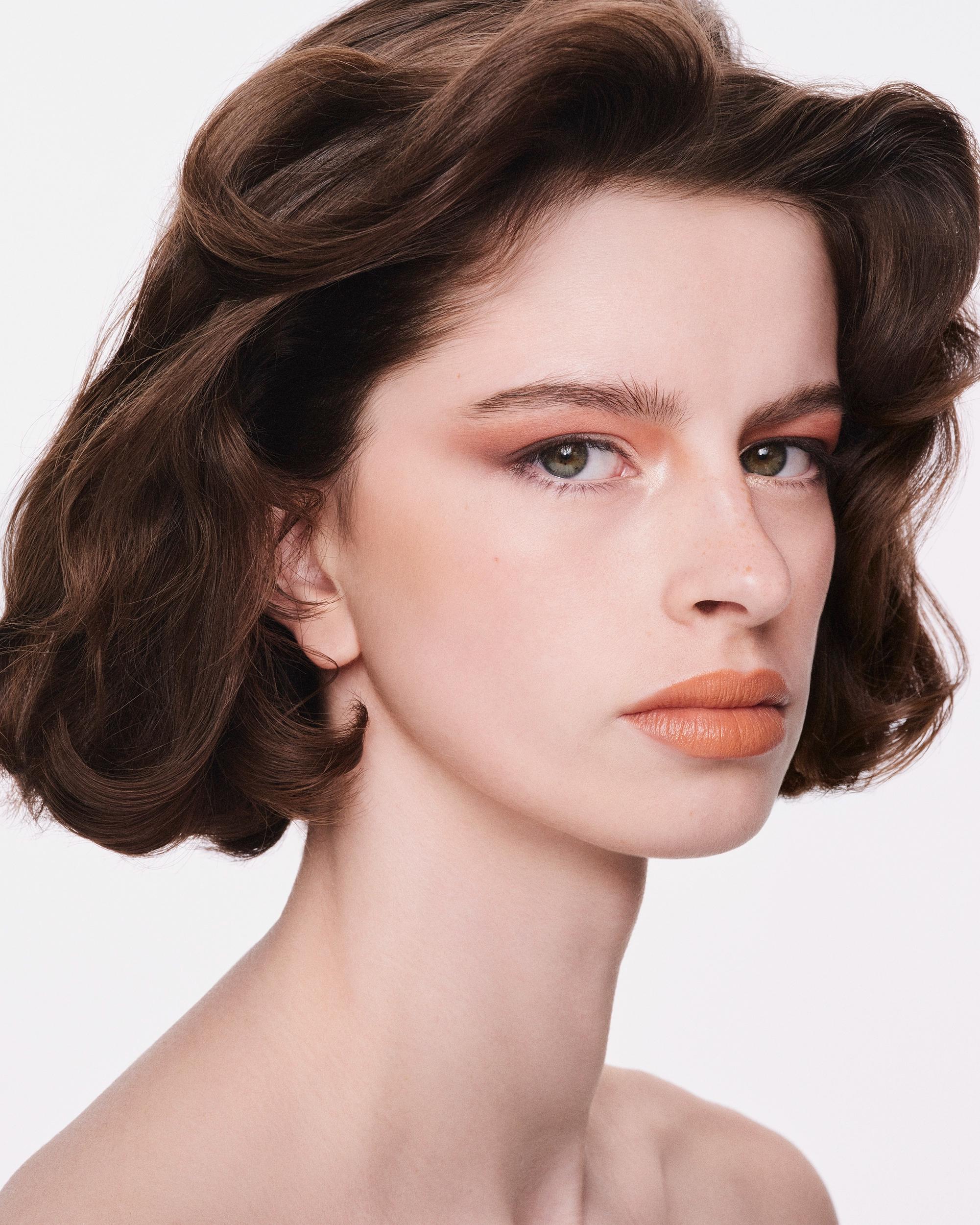 Portrait of a woman with voluminous brown vintage waves, green eyes, terracotta eyeshadow, and matte apricot lips, looking over her shoulder at the camera.