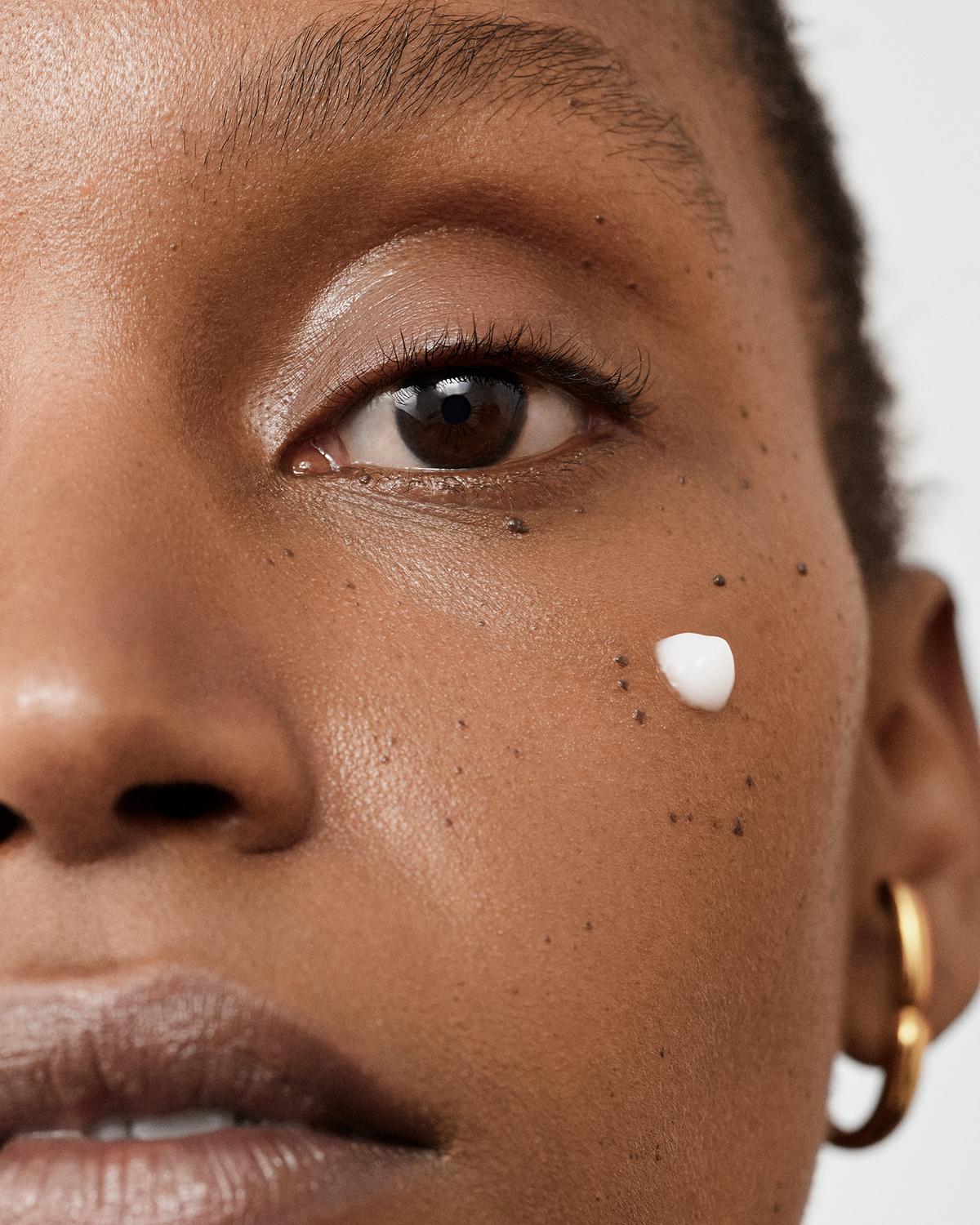 Close-up of a woman's face with glowing skin and a small dab of white cream on her cheek.