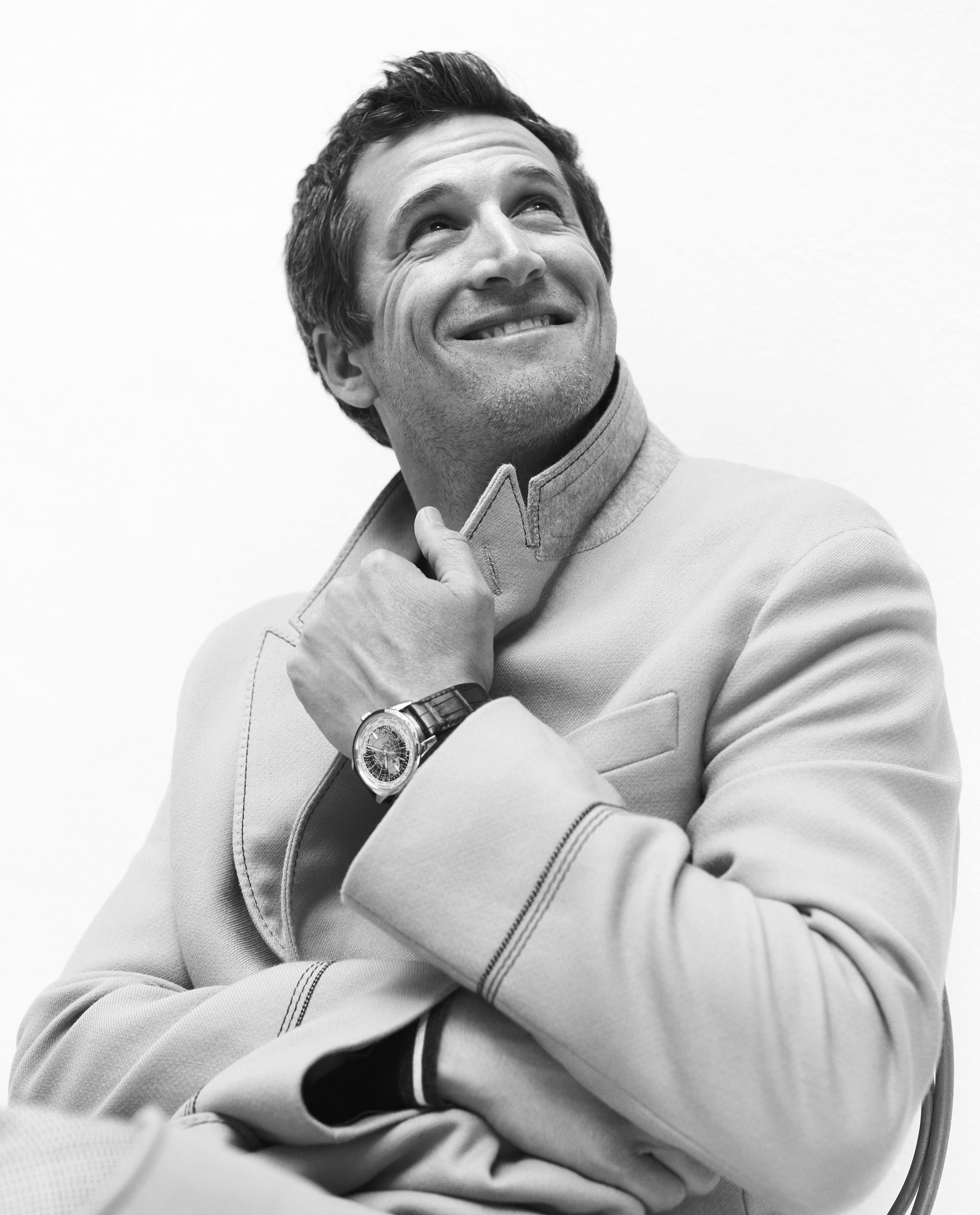 Black-and-white portrait of French actor Guillaume Canet smiling and looking upward in a light tailored jacket, a wristwatch on the hand at his collar