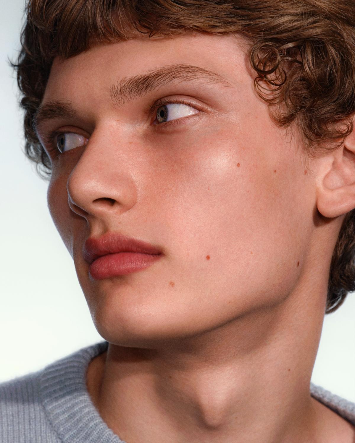 Side-profile close-up of a young man with curly brown hair and blunt bangs, glowing skin, and rosy-mauve lips, wearing a gray knit sweater and glancing sideways