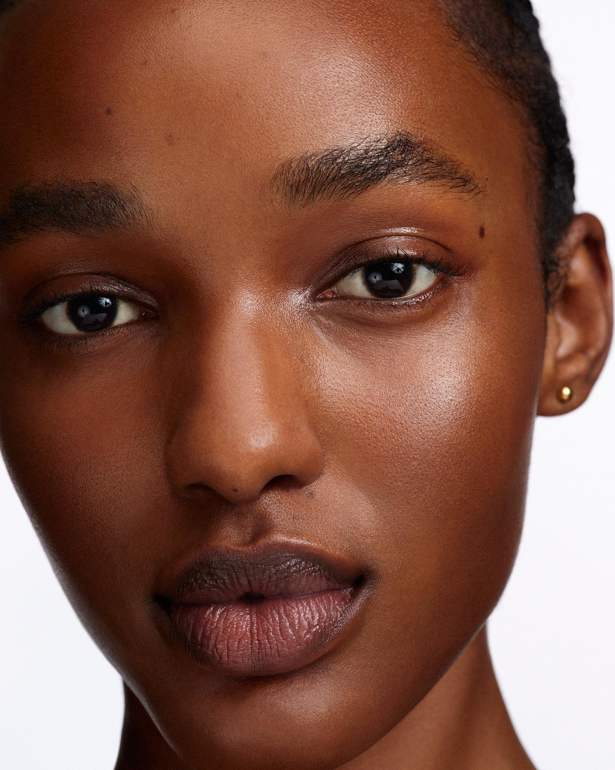 Close-up portrait of a woman with radiant deep-toned skin, dark brown eyes, and muted berry-brown lips, wearing a small gold stud earring.