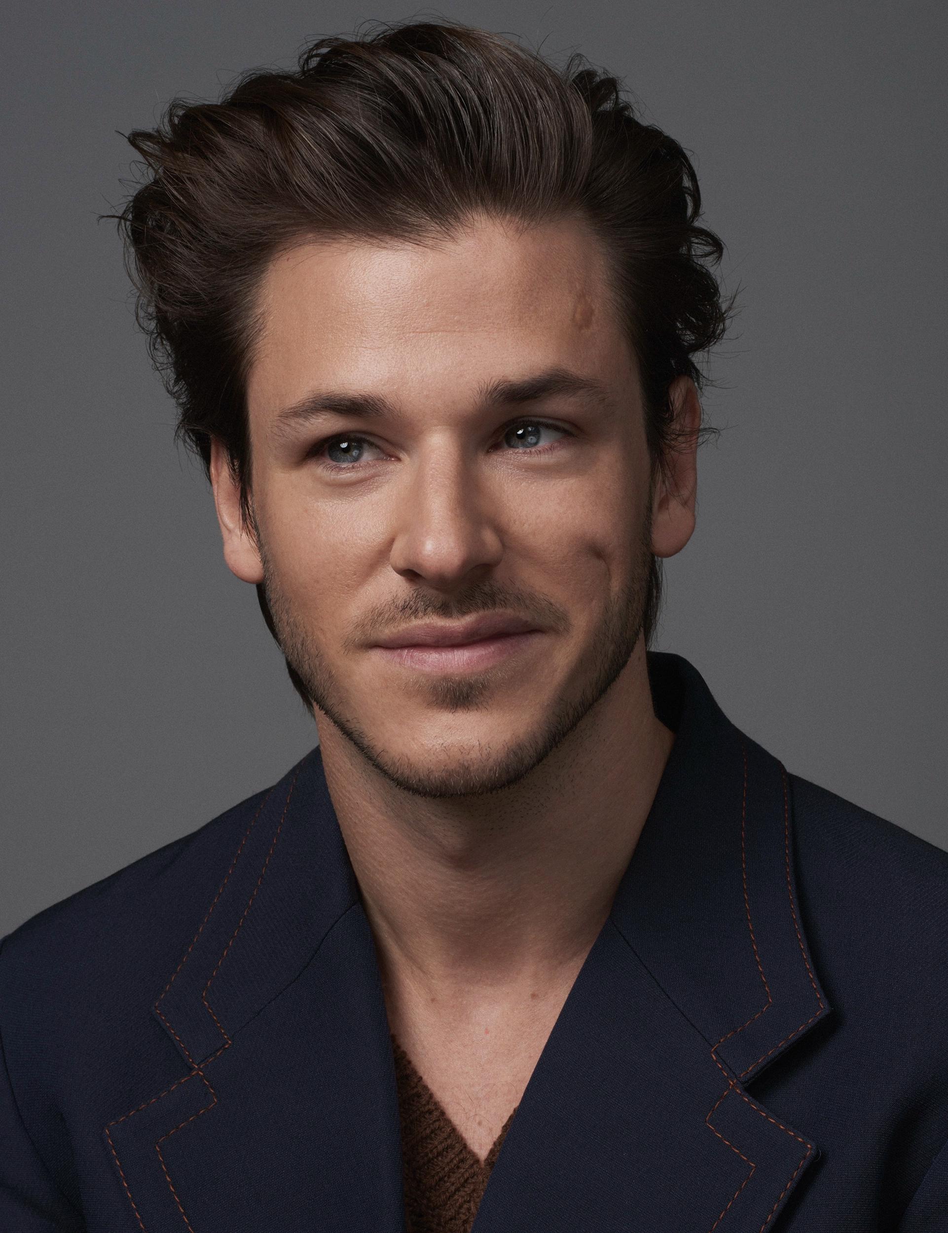 Portrait of actor Gaspard Ulliel with swept-back dark hair and light stubble, smiling slightly in a navy jacket with orange topstitching over a brown knit