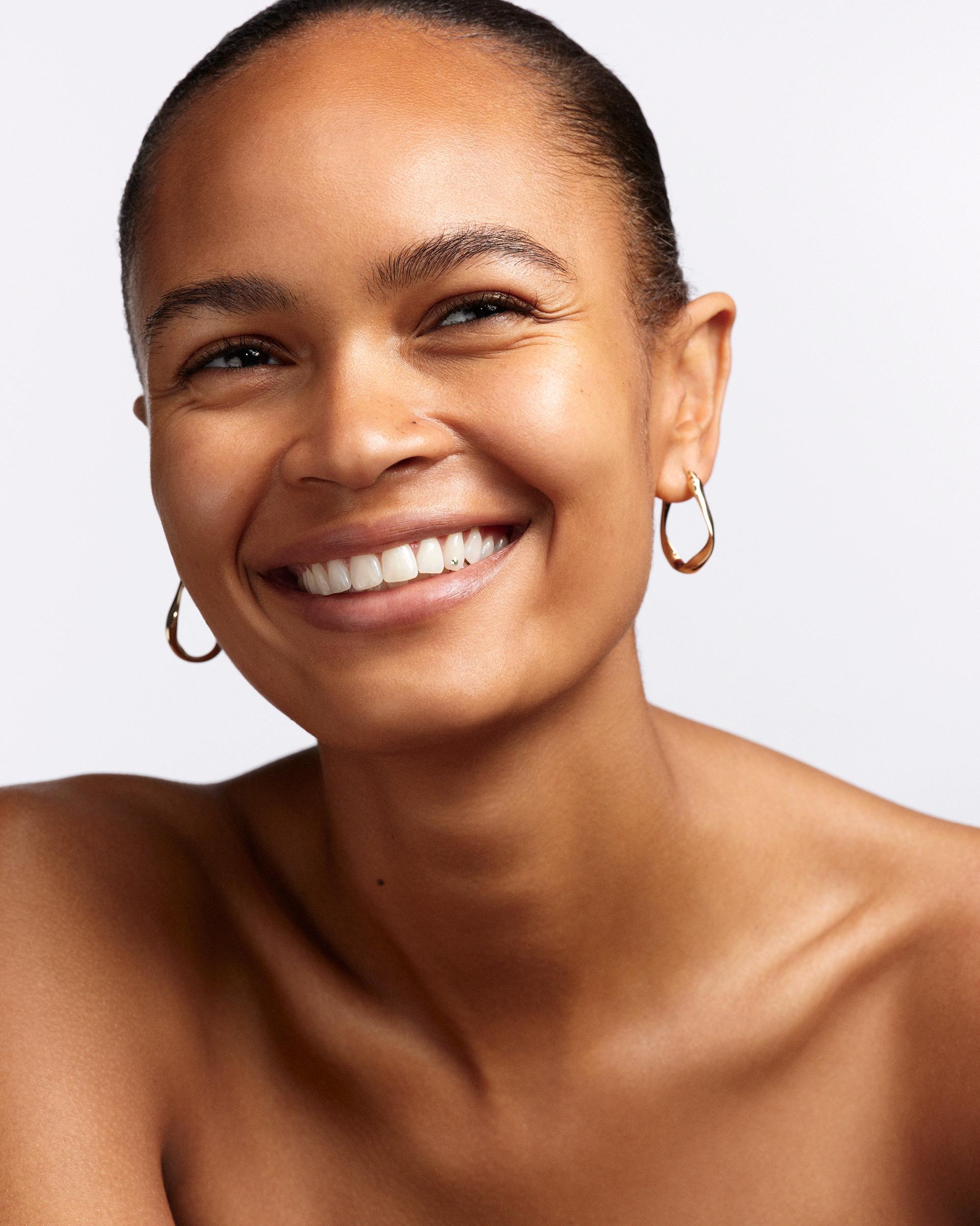 Smiling woman with bare shoulders, glowing skin, slicked-back hair, and sculptural gold hoop earrings, looking up and away from the camera.