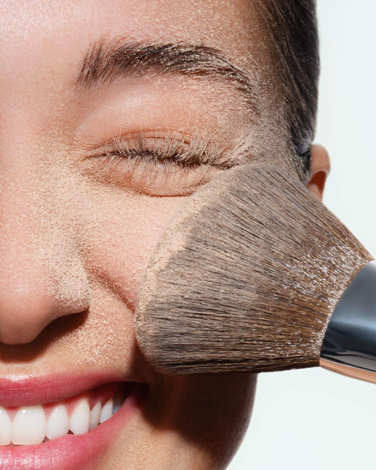 Close-up of a smiling woman with closed eyes as a large fluffy brush sweeps loose beige powder across her cheek, dusting her skin and brow.