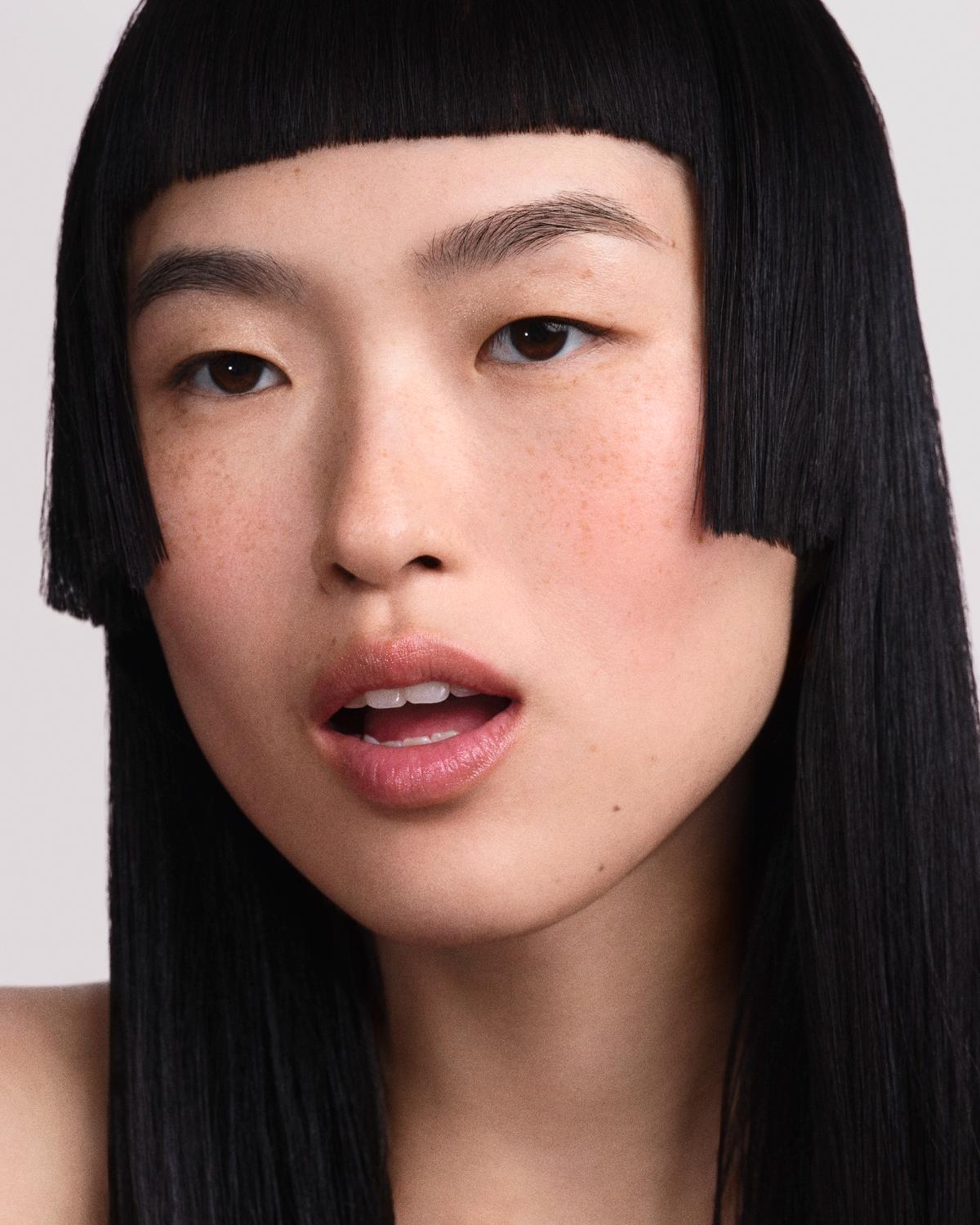 Portrait of a woman with long, jet-black hair and sharp geometric bangs, light freckles, and soft pink lips, looking at the camera.