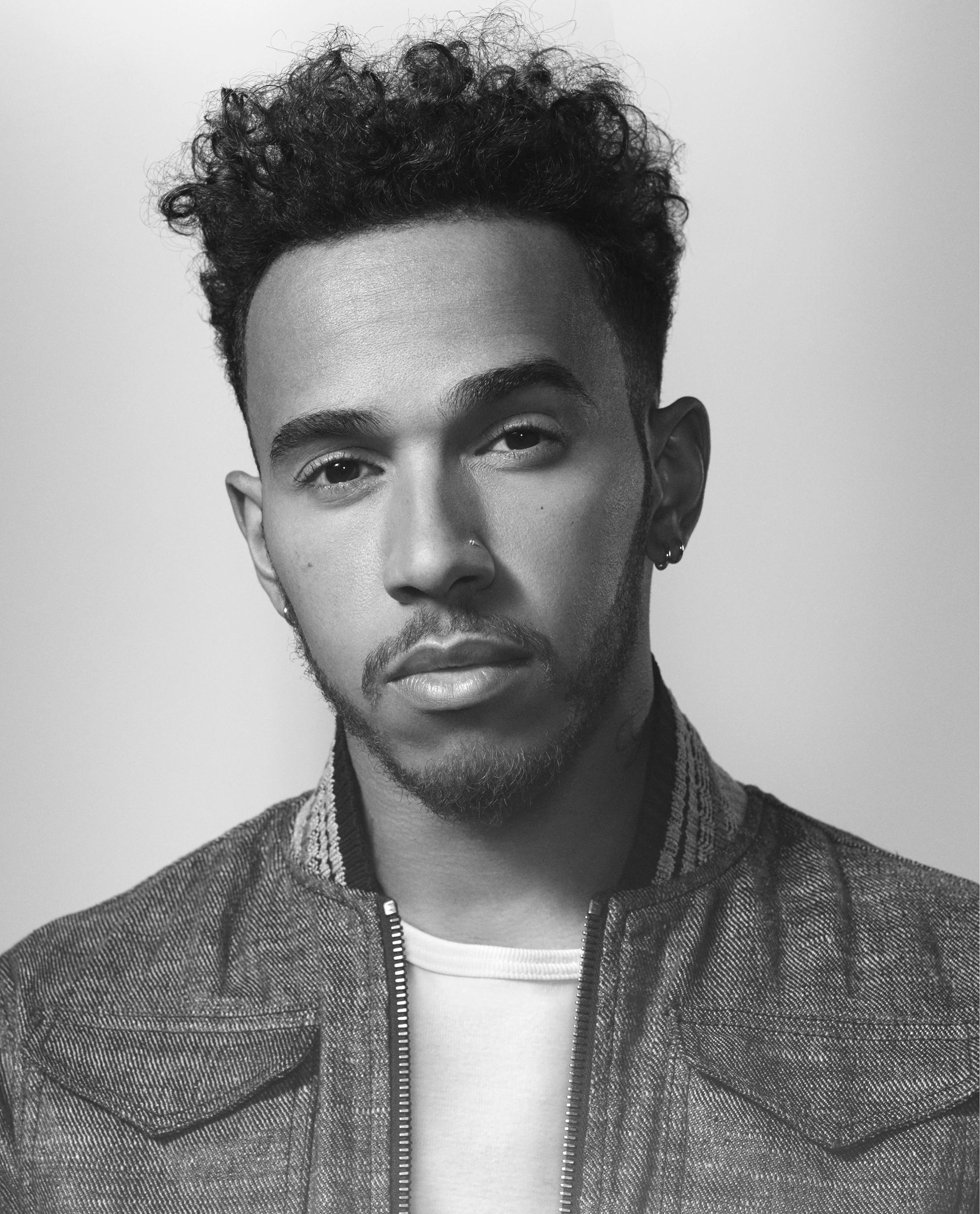 Black-and-white portrait of Lewis Hamilton with curly hair, a goatee, hoop earrings, and a nose stud, in a textured zip-up jacket over a white tee