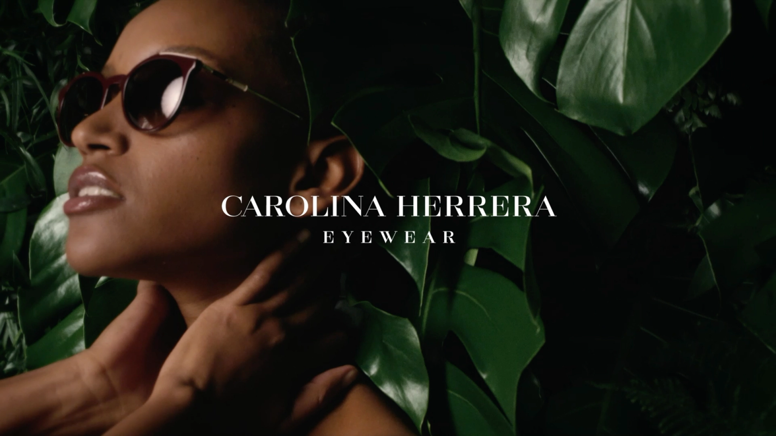 Carolina Herrera Eyewear campaign video still: a woman in round burgundy sunglasses tilts her head back among glossy green leaves, a hand at her neck.