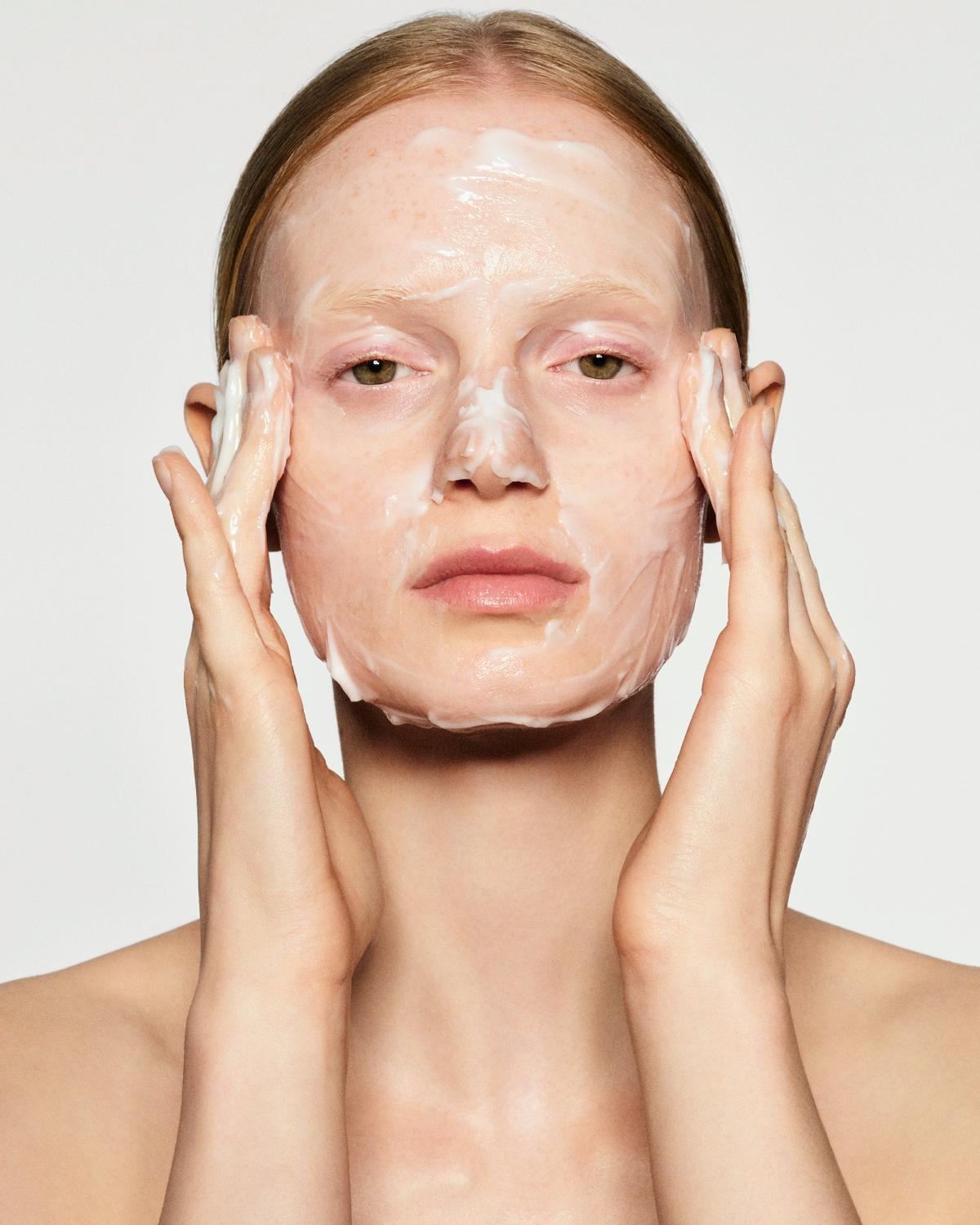 Woman with slicked-back hair massaging a thick white cream or mask over her face, with both hands pressed to her cheeks.