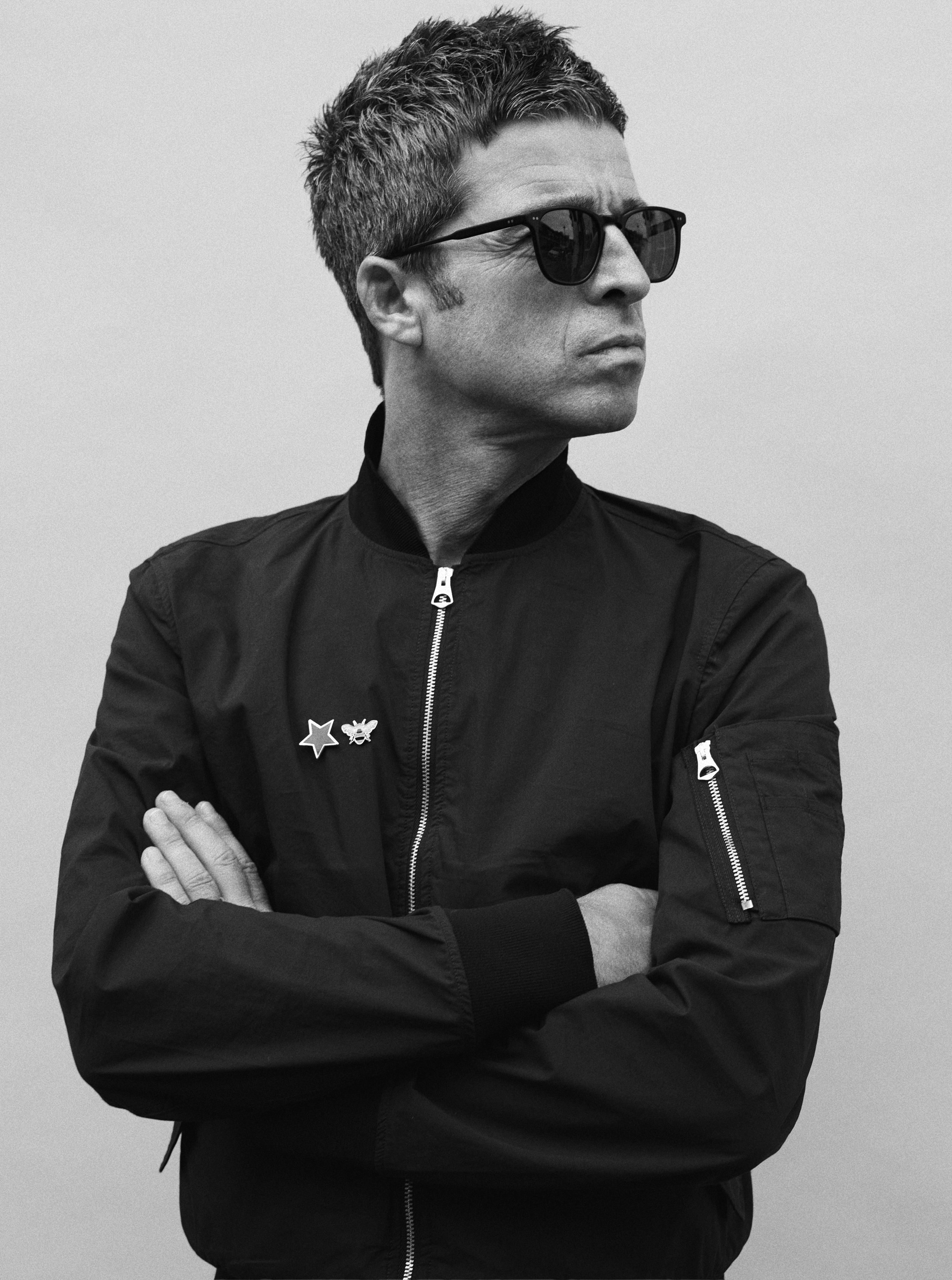 Black-and-white portrait of musician Noel Gallagher in sunglasses and a black zip-up bomber jacket with star and bee pins, arms crossed, looking off to the side