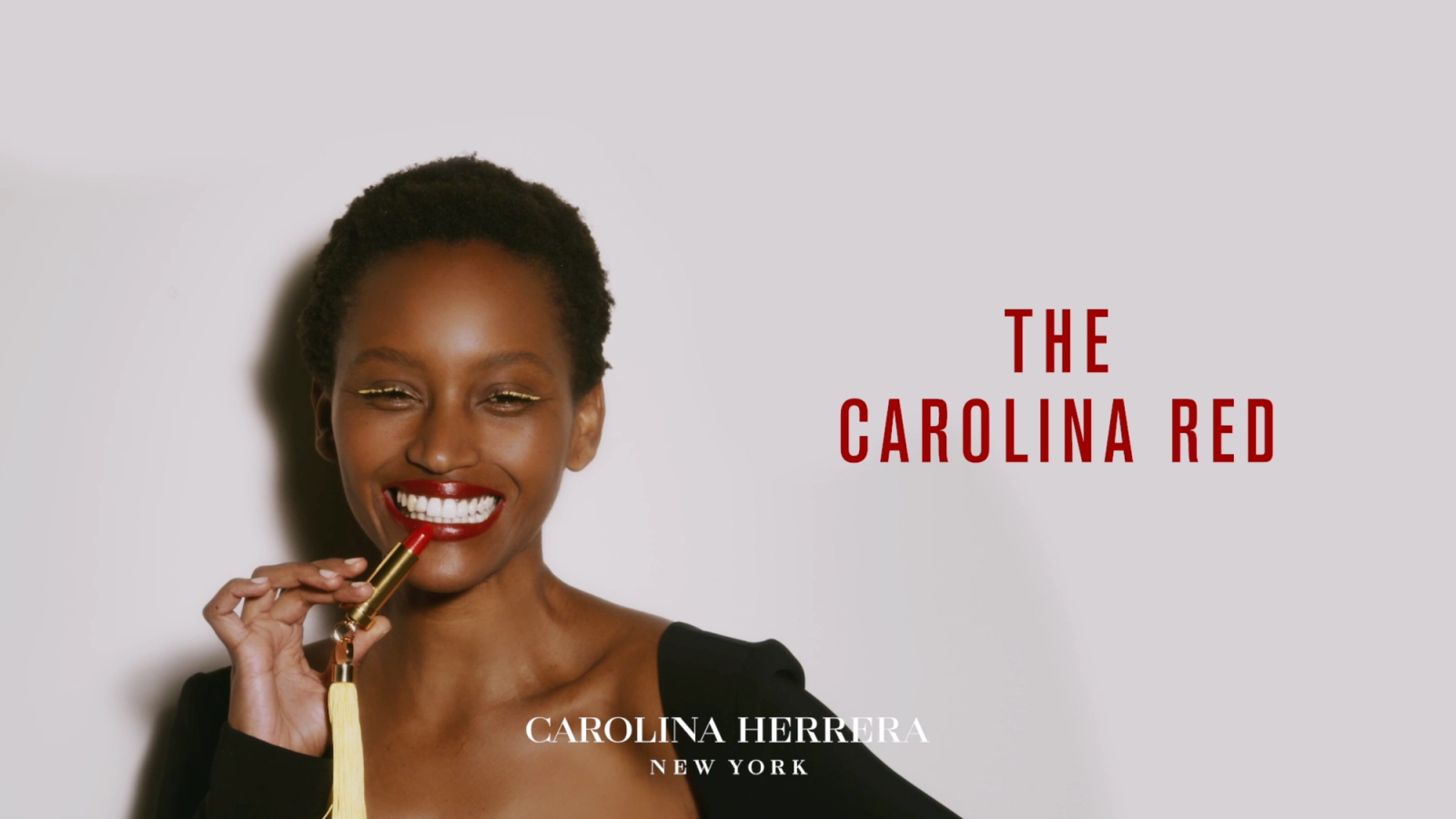 Carolina Herrera campaign video still: a smiling woman with yellow eyeliner and red lips holds a gold tasseled lipstick beside the text “The Carolina Red.”