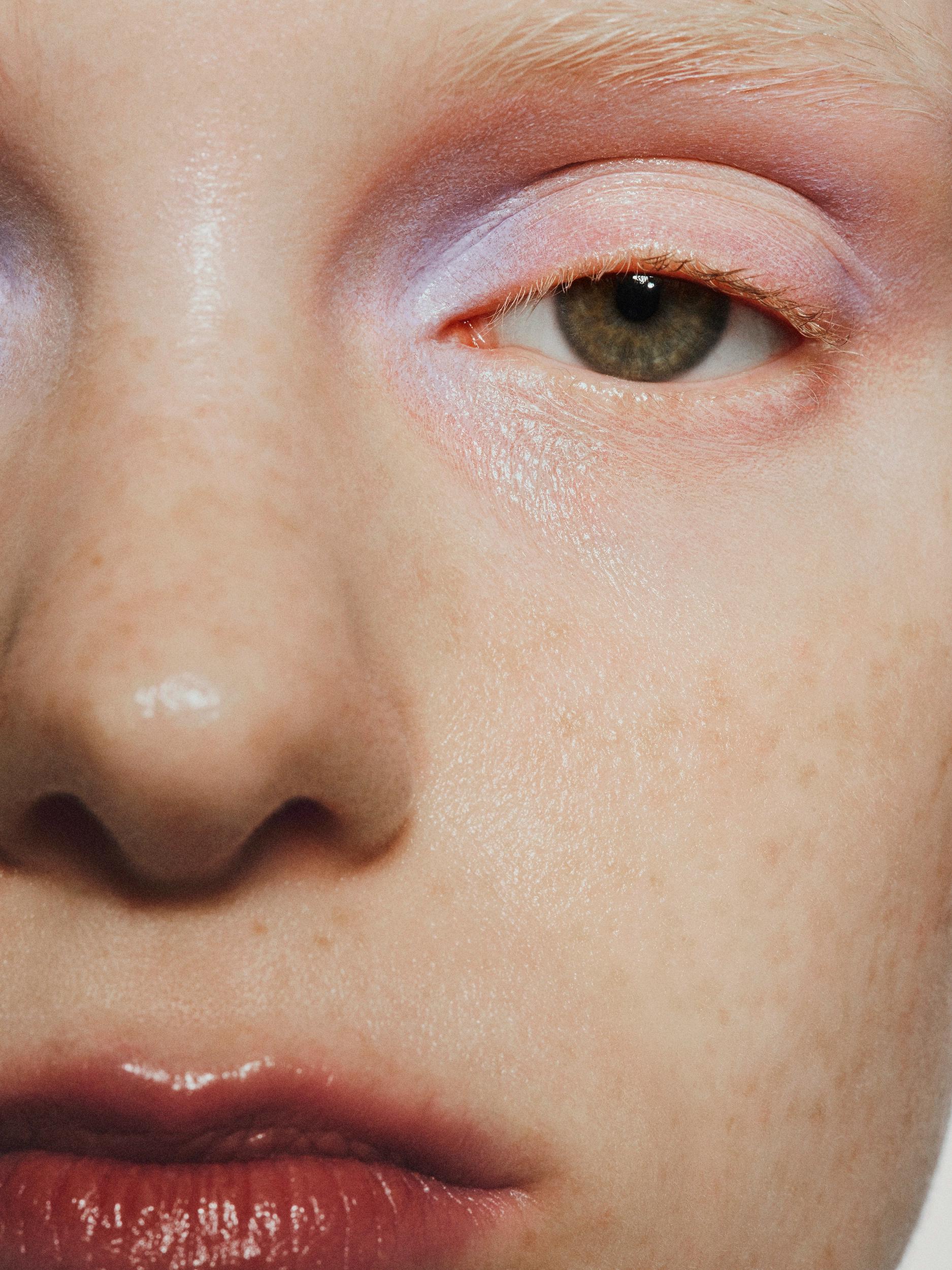 Extreme close-up of half a face with freckled skin, a green eye with lilac and pink eyeshadow, and glossy berry-tinted lips.