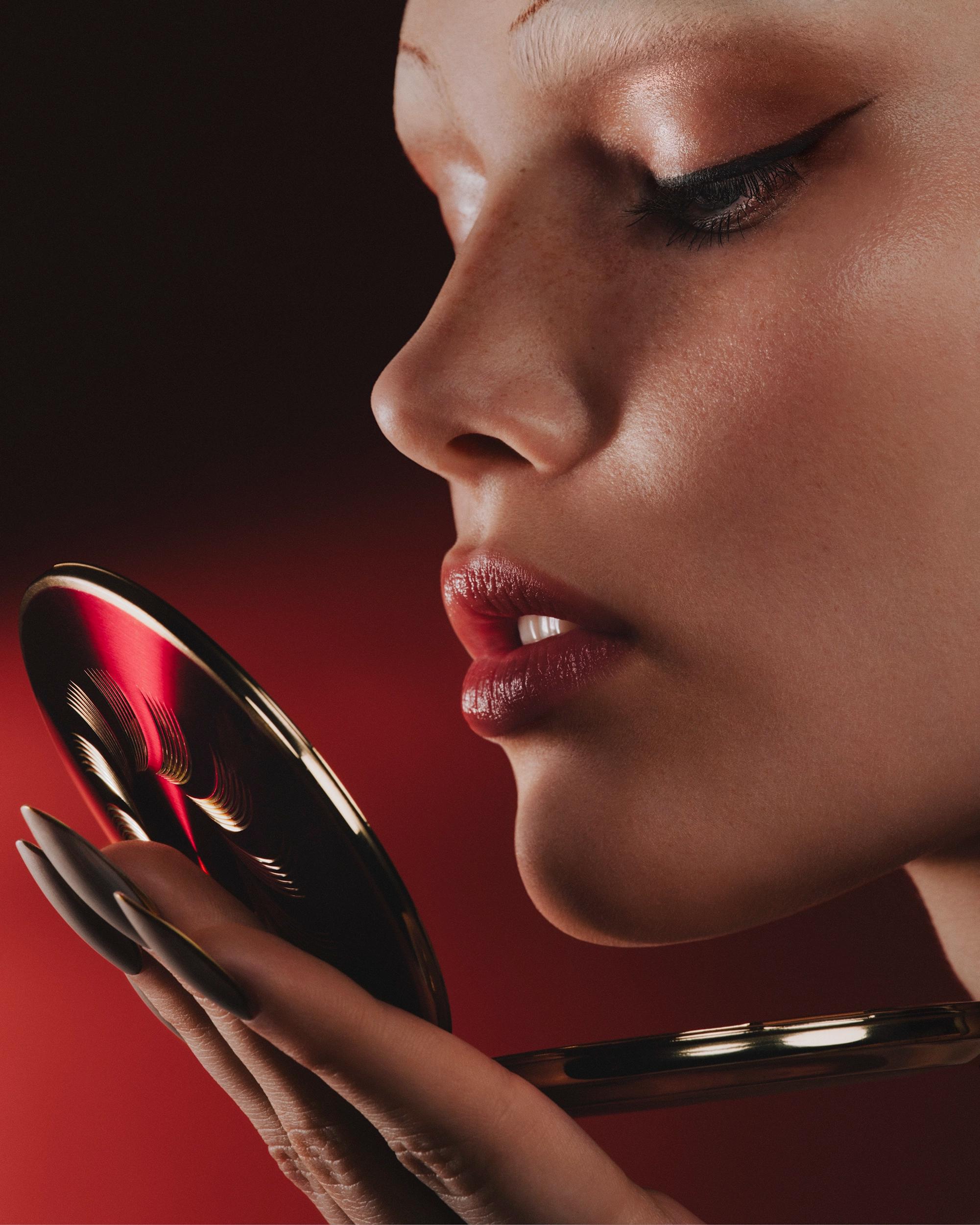 Side-profile close-up of a woman with winged eyeliner and glossy deep red lips, holding a gold compact mirror in long pointed nails against a red backdrop.