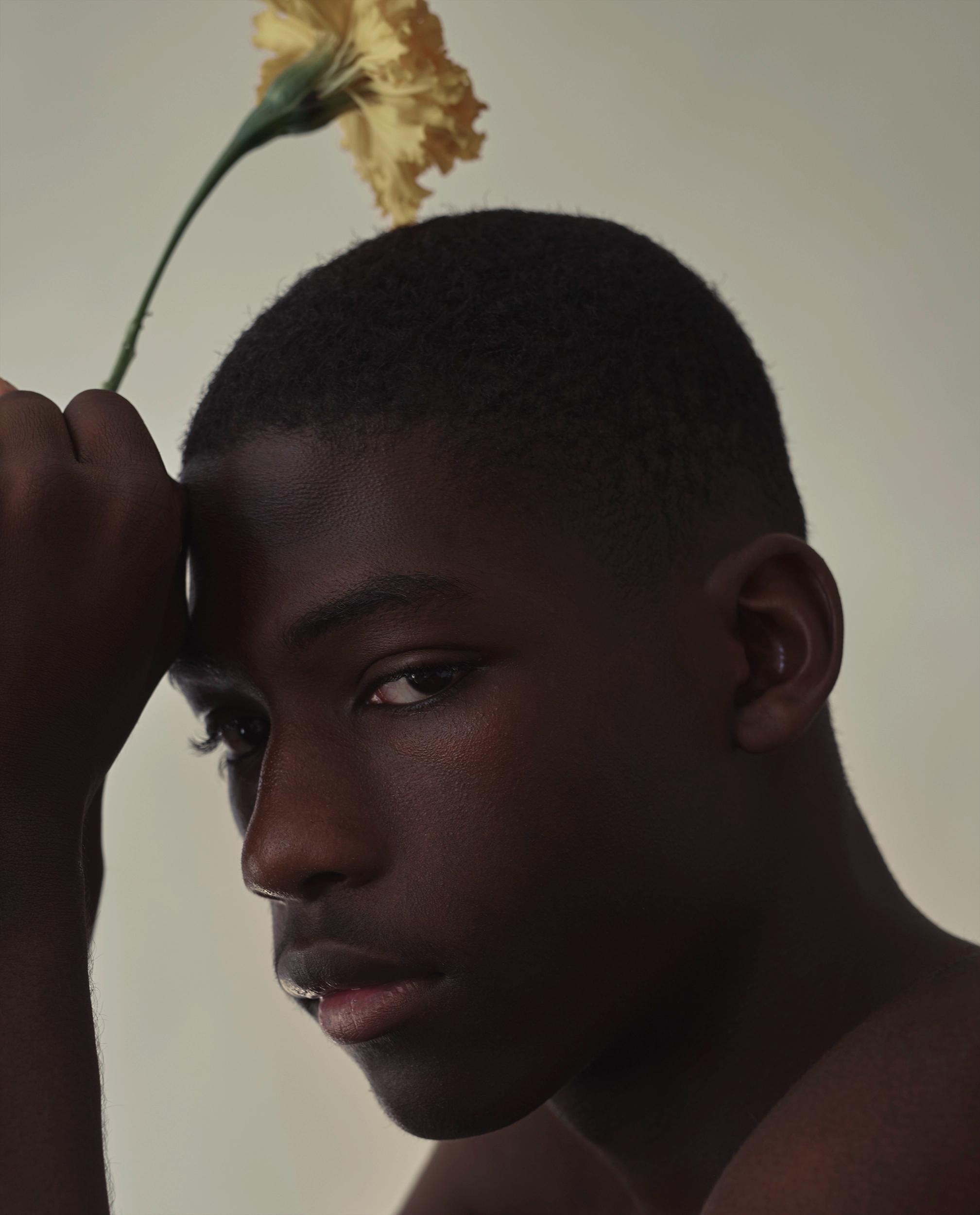 Moody portrait of a young man with a buzz cut and bare shoulders, his fist against his forehead and eyes cast down, with a yellow carnation above his head