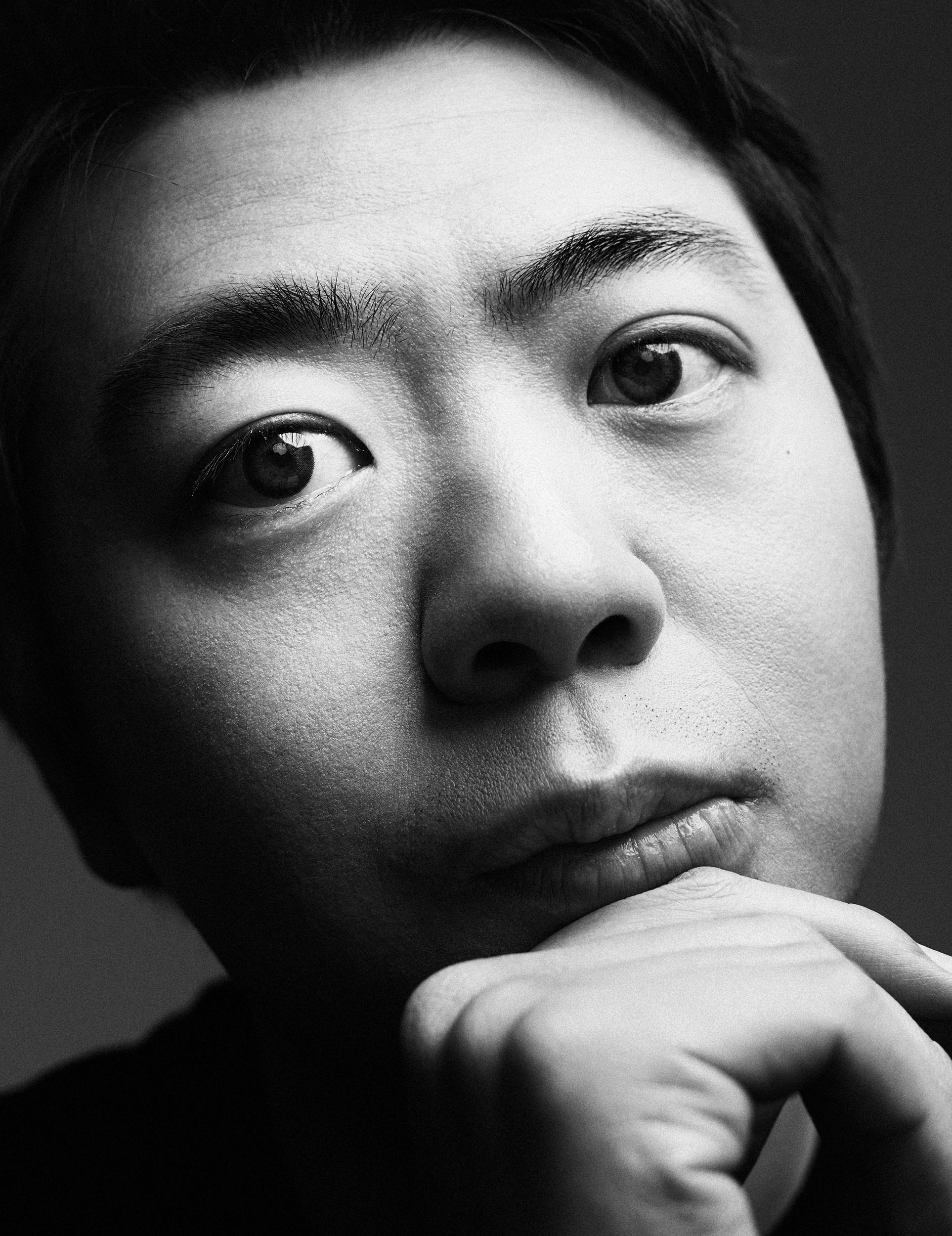 Black-and-white close-up portrait of pianist Lang Lang resting his chin on his hand and gazing intently at the camera against a dark background.