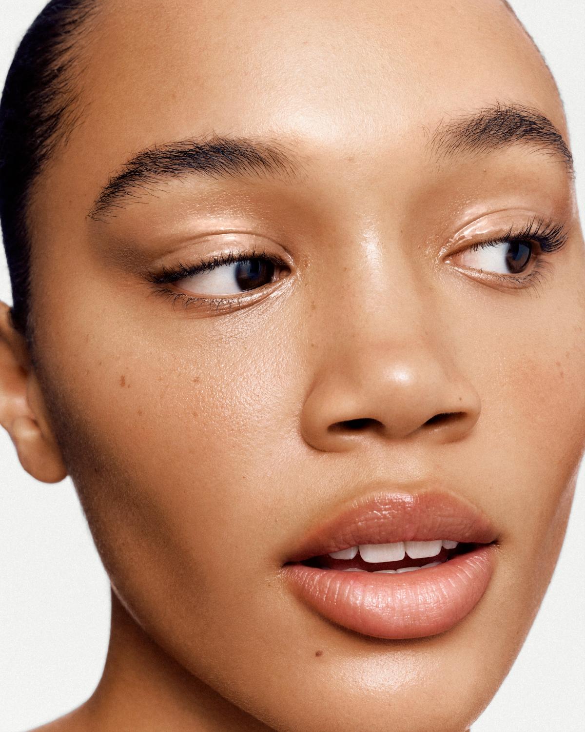 Close-up beauty portrait of a woman glancing to the side, with dewy skin, glossy shimmer on her eyelids, and soft nude lips