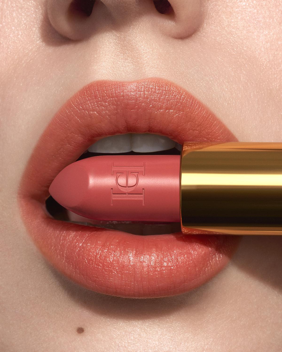 Close-up of coral-peach lips holding a rosy-pink lipstick with an embossed monogram in a gold tube.