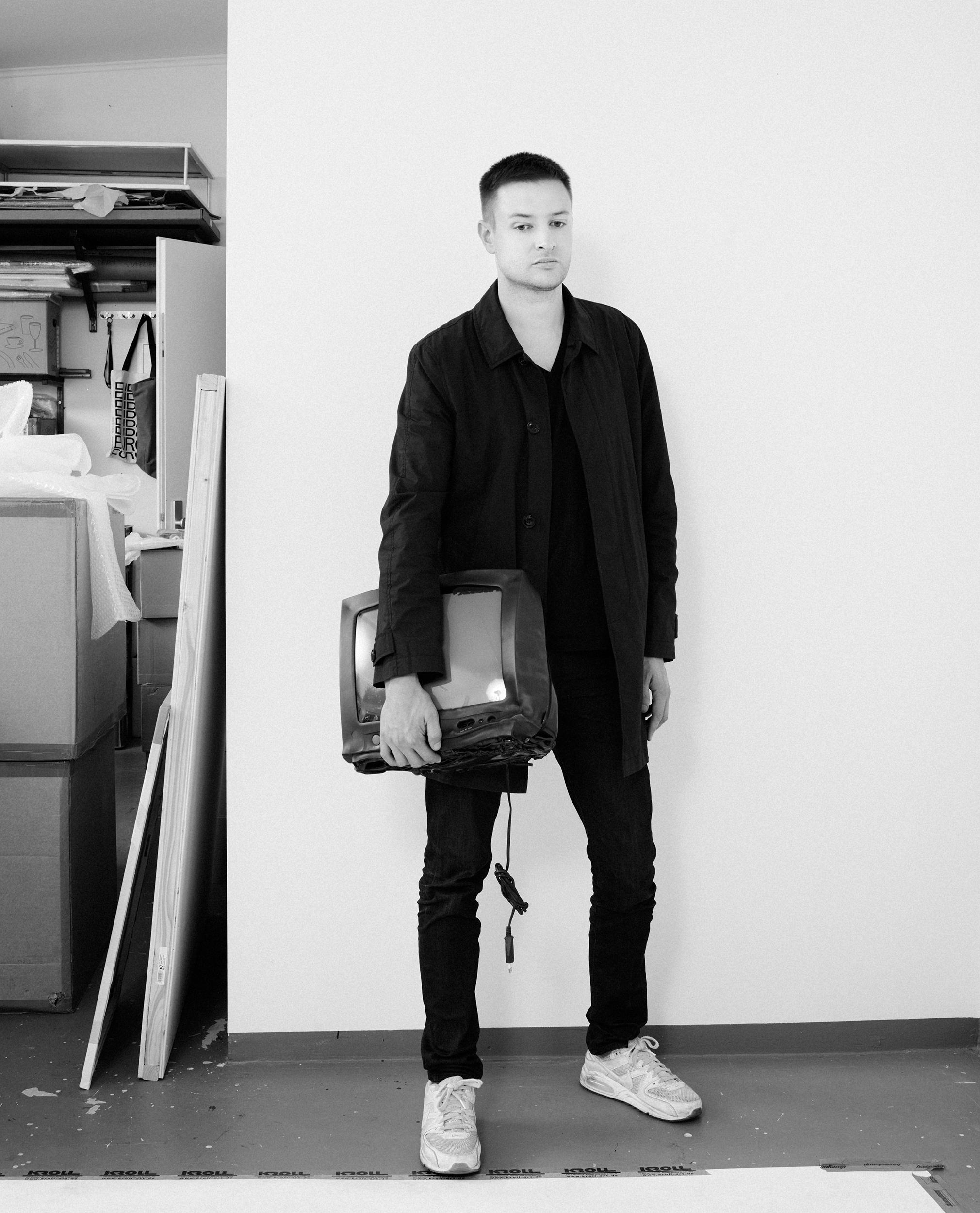 Black-and-white full-length portrait of artist Simon Denny in a dark coat and white sneakers, holding an old CRT television in a cluttered studio.