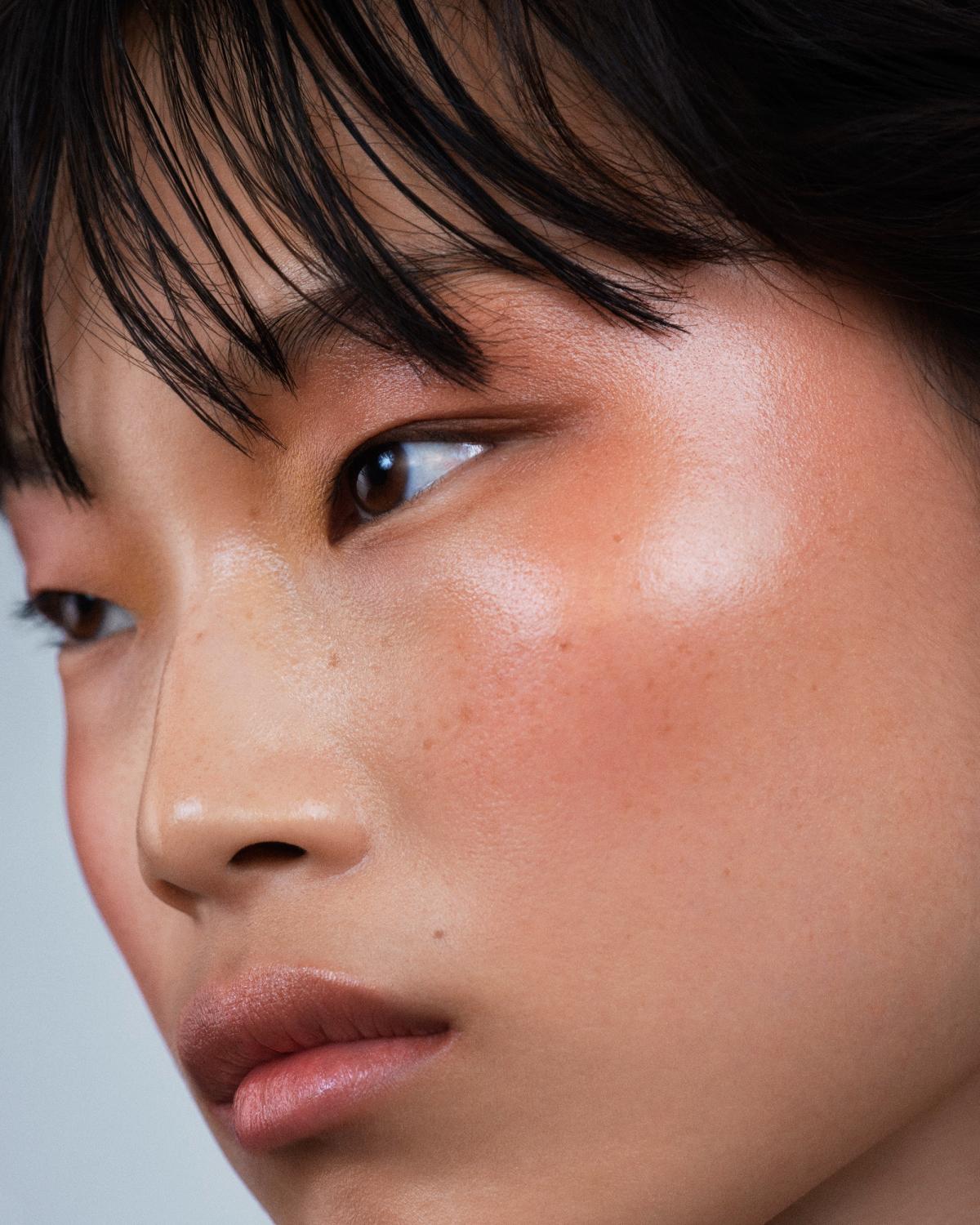 Close-up of a woman with wispy black bangs glancing down and to the side, with warm peach blush, a dewy highlight on her cheekbone, and soft mauve-nude lips.