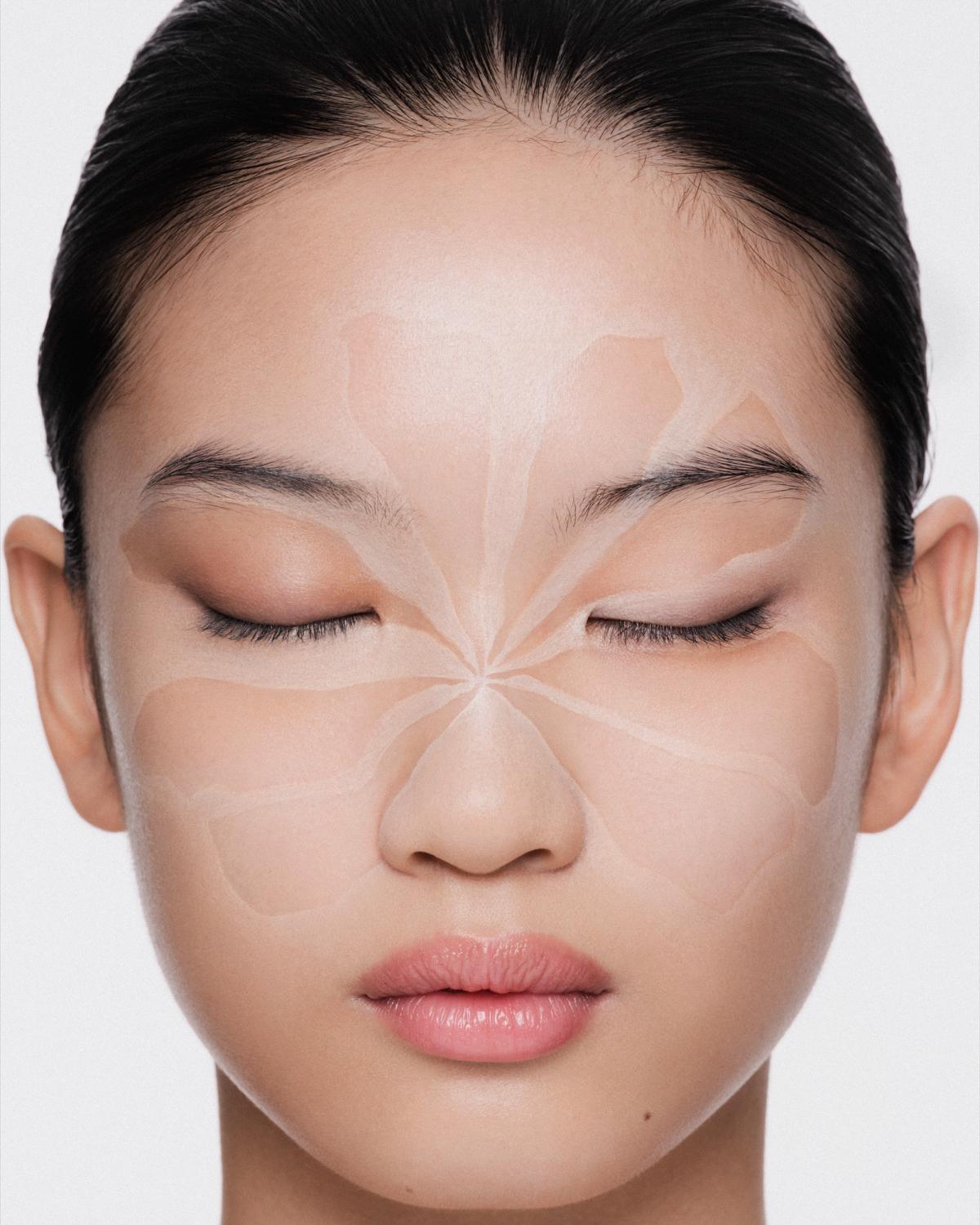 Close-up of a woman with closed eyes and slicked-back hair, a flower-petal design painted across her face in nude and ivory tones, and soft pink lips.
