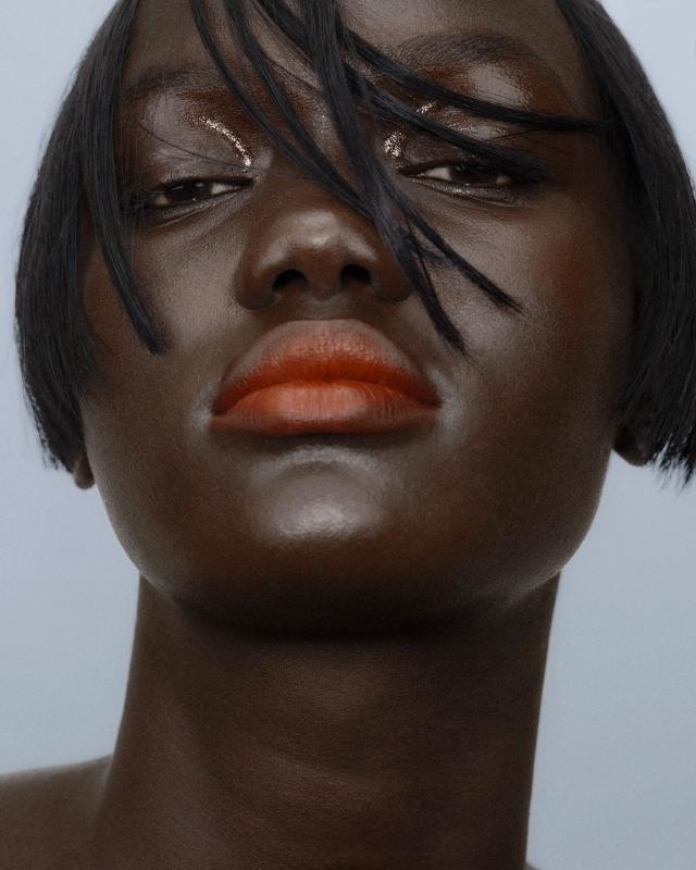 Low-angle close-up of a woman with a sleek black bob and strands falling across her face, glossy skin, a pearly highlight on her chin, and burnt-orange lips