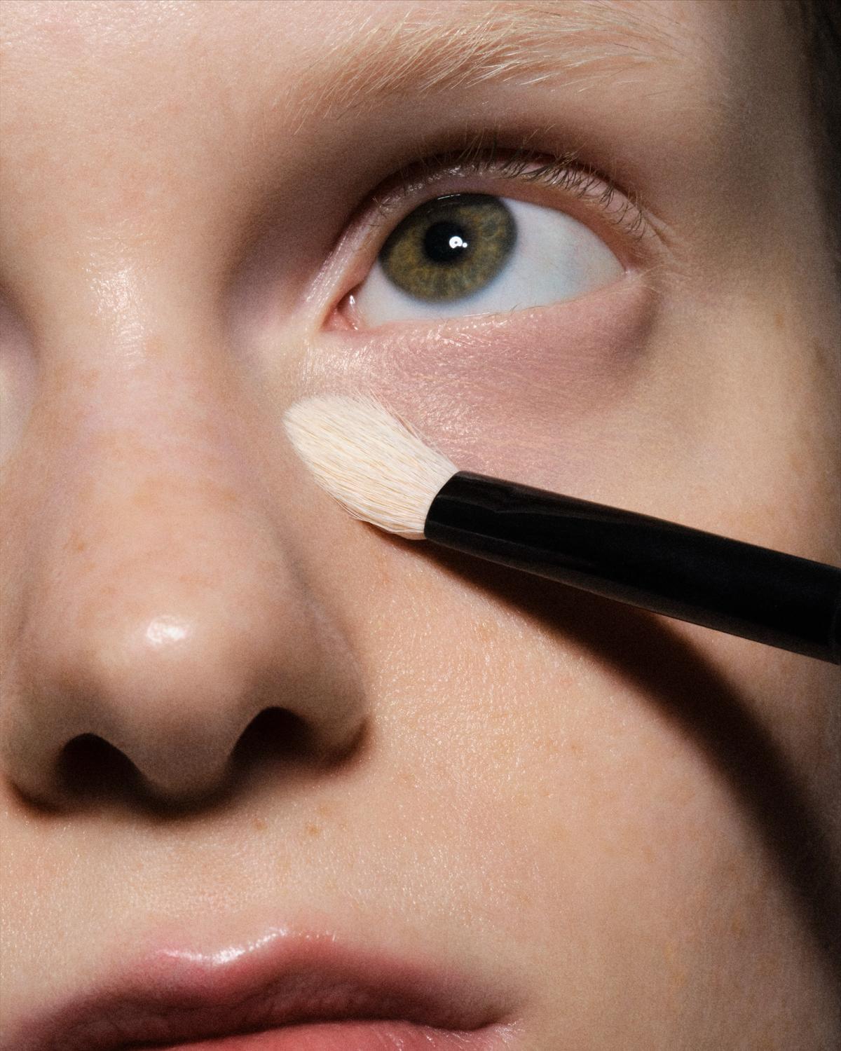 Extreme close-up of a green eye and bleached brow as a fluffy white brush dabs cream concealer under the eye on freckled, dewy skin