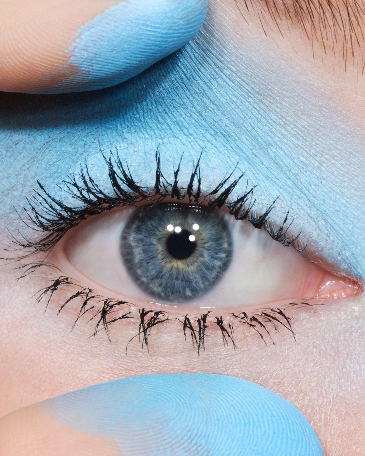 Extreme close-up of a gray-blue eye with clumpy black lashes, powder-blue eyeshadow on the lid, and two blue-dusted fingertips framing the eye.