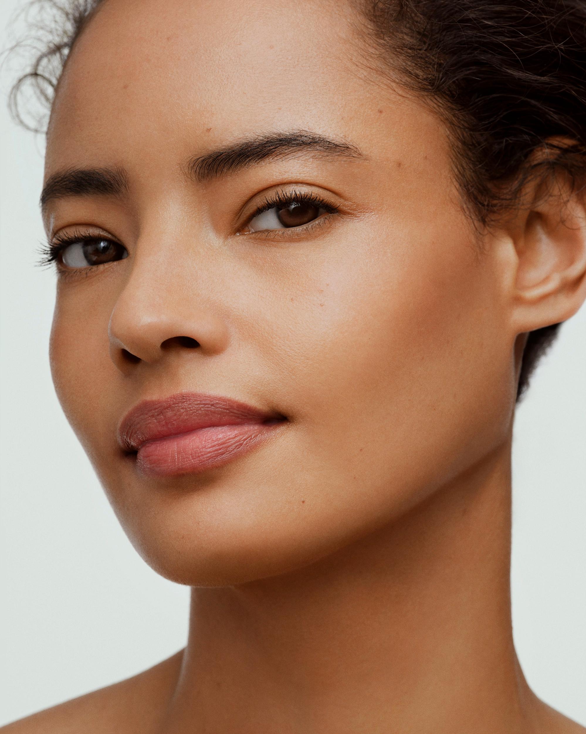Close-up beauty portrait of a woman with natural, glowing skin, defined brows, and soft rose-nude lips, her dark hair pulled up, glancing to the side