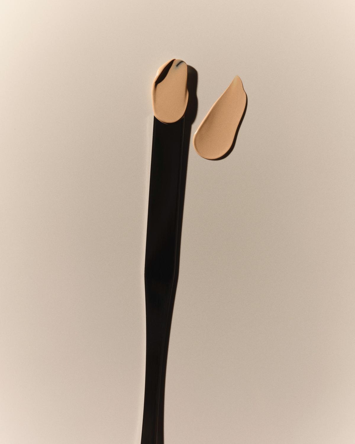Two glossy dabs of creamy beige product, one sitting on the tip of a long, slim black spatula, the other beside it, on a soft neutral background