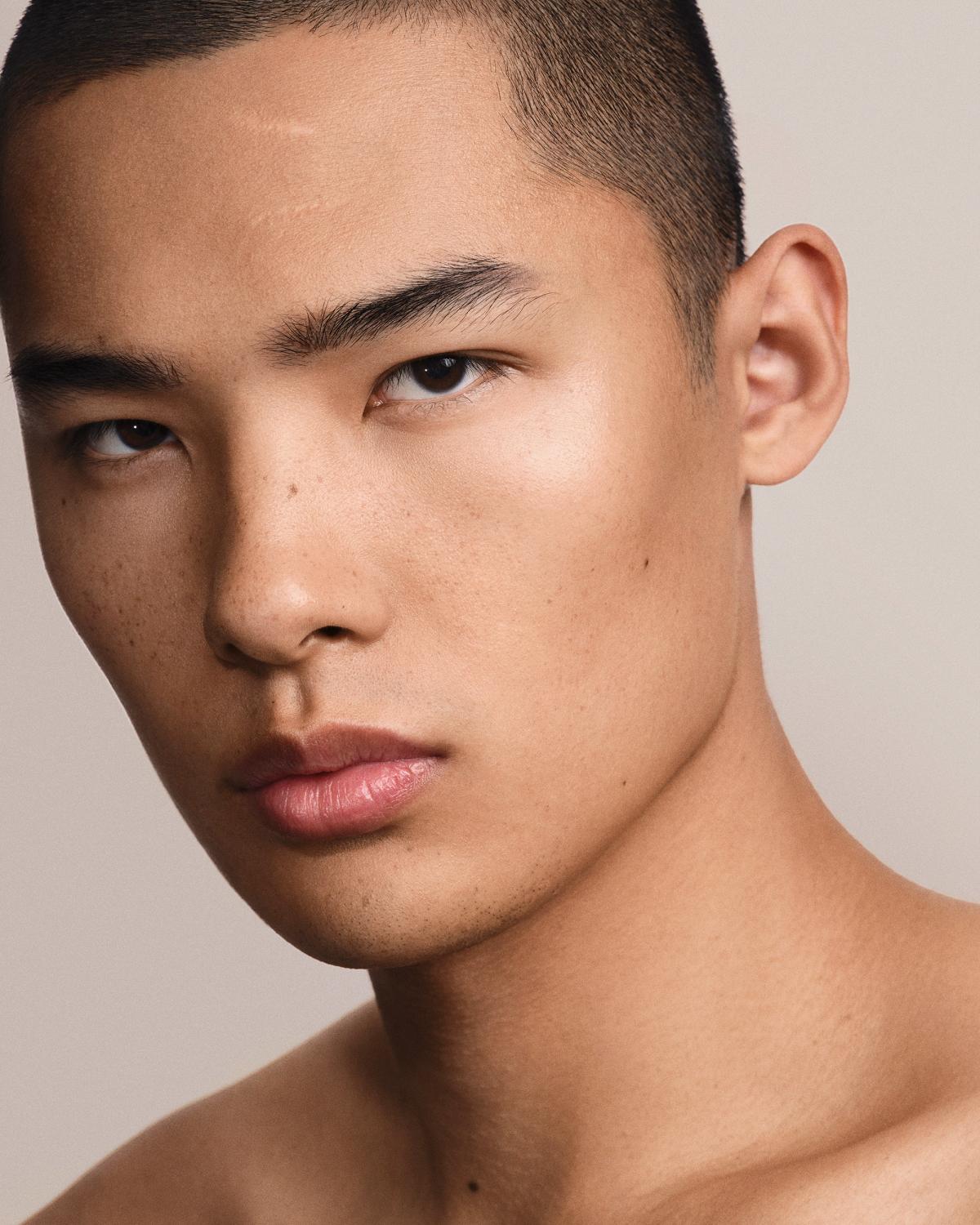 Close-up portrait of a young man with a buzz cut, light freckles, glowing skin, and glossy pink lips, with bare shoulders, looking intently at the camera.