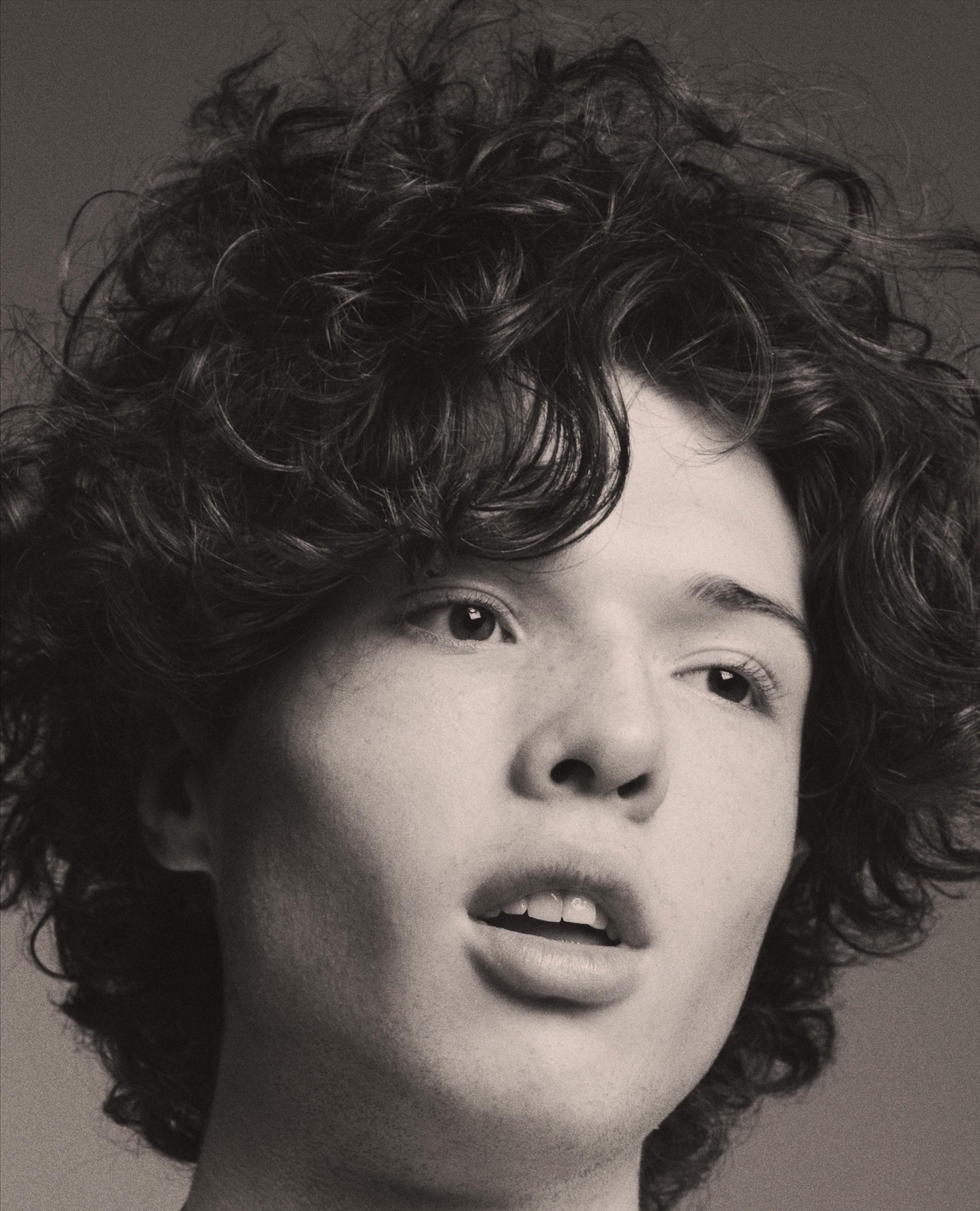 Sepia-toned close-up portrait of a young person with voluminous dark curls falling over their forehead, lips slightly parted, gazing upward past the camera