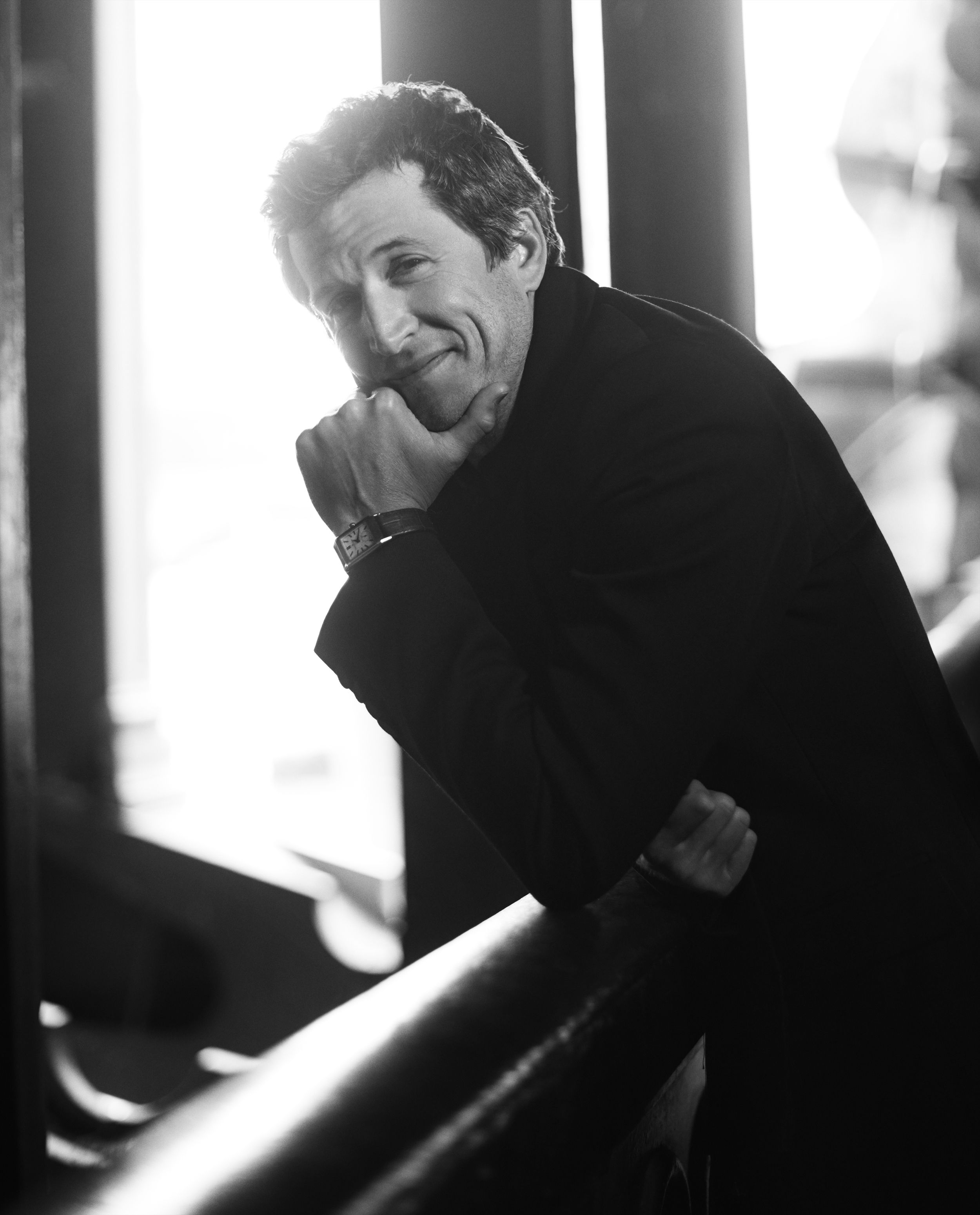 Black-and-white portrait of actor Guillaume Canet leaning on a dark railing in a black coat, his chin resting on his fist and a rectangular watch on his wrist