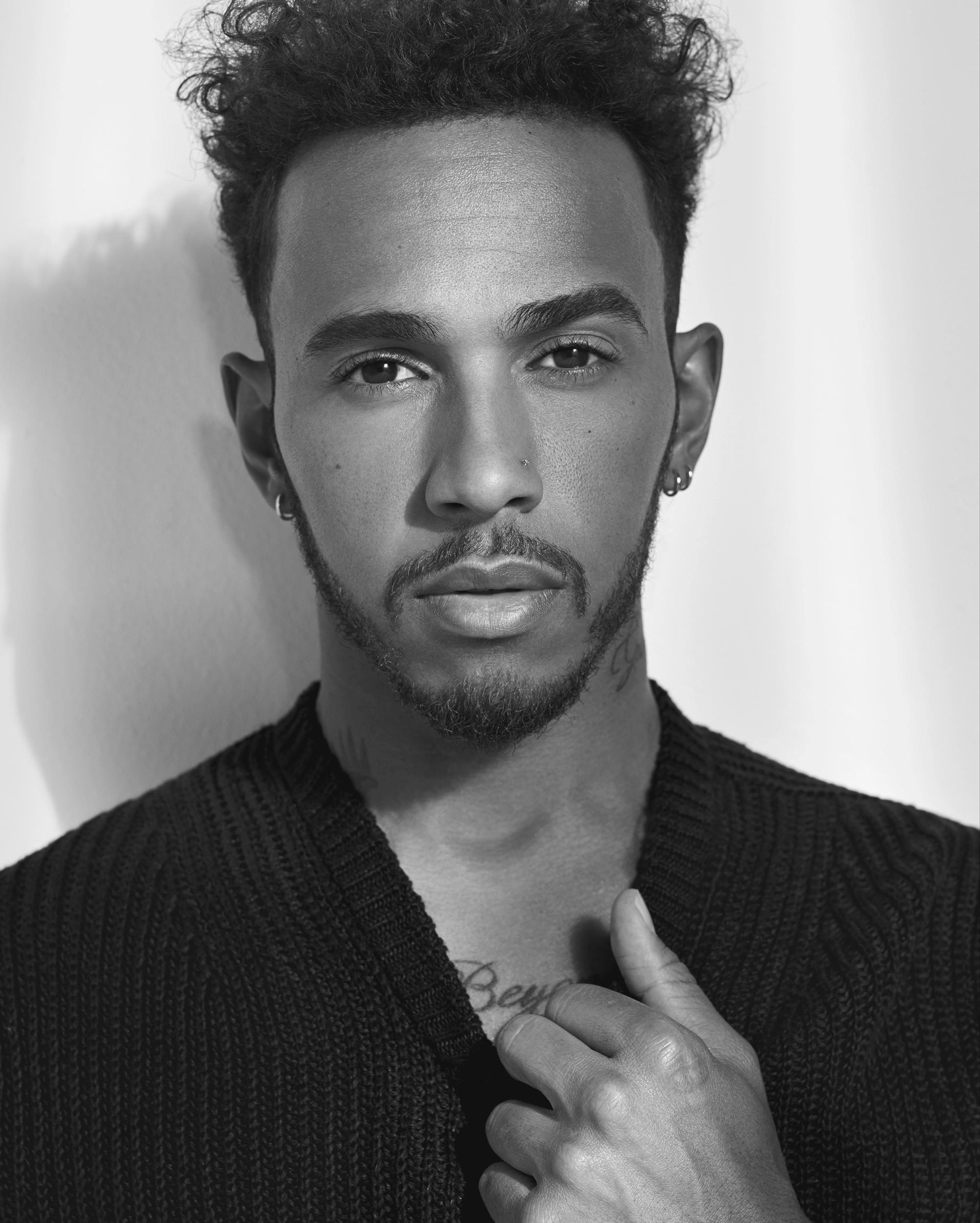 Black-and-white portrait of F1 driver Lewis Hamilton in a dark knit cardigan, with curly hair and hoop earrings, hand at his collar, gazing at the camera.