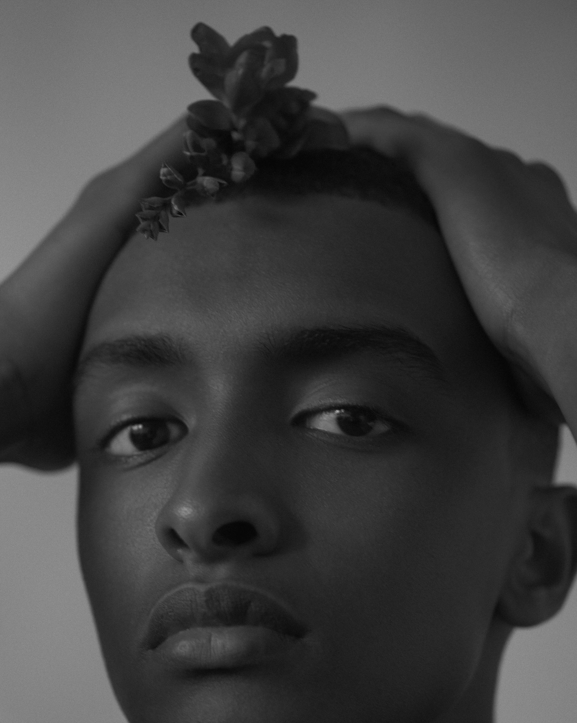 Moody black-and-white close-up of a young man looking at the camera with both hands on his head and a sprig of small flowers resting in his hair