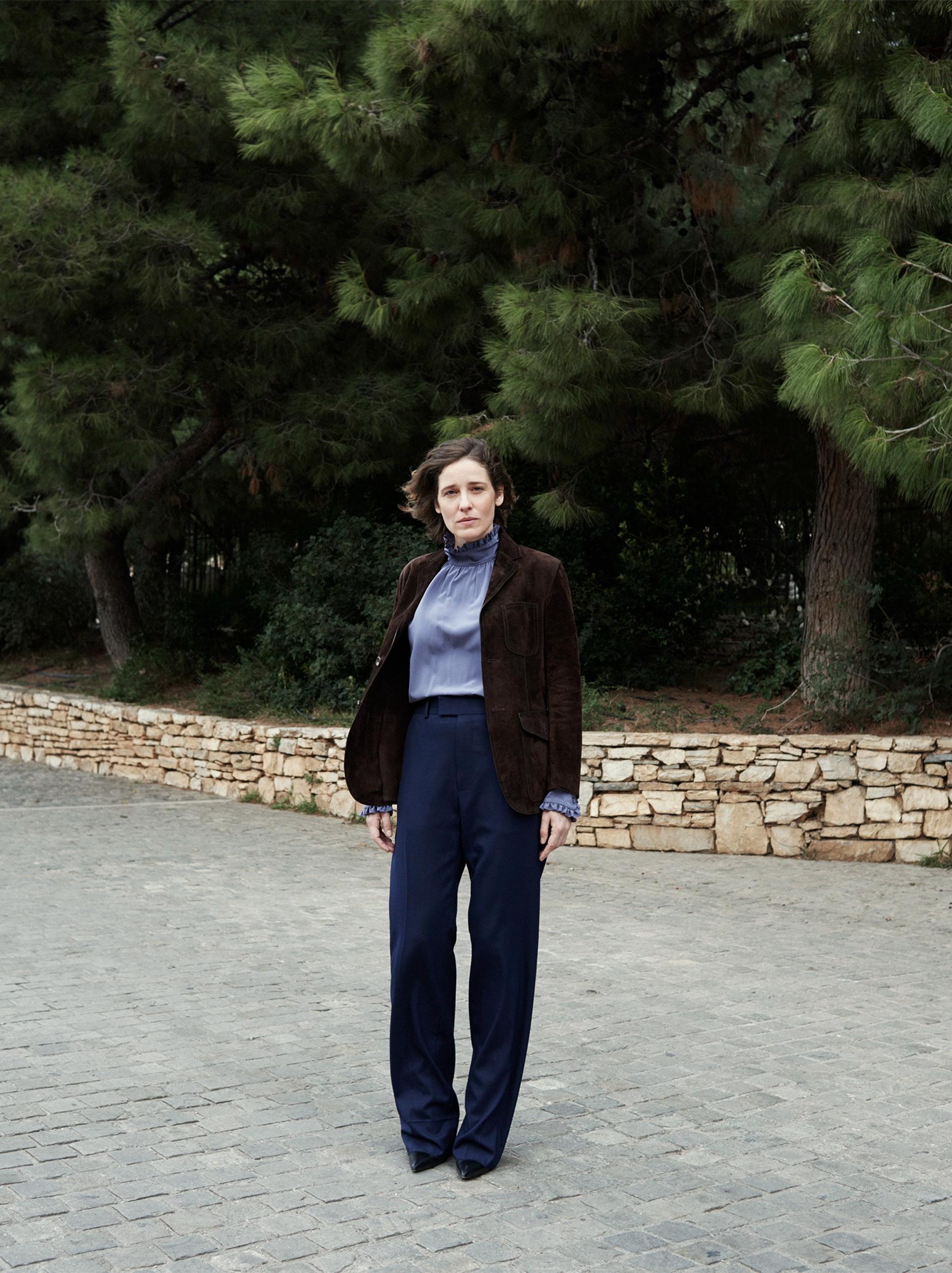 Actress Angeliki Papoulia standing on a cobblestone path in a brown suede jacket, periwinkle ruffled blouse, and navy trousers, with pine trees behind her.
