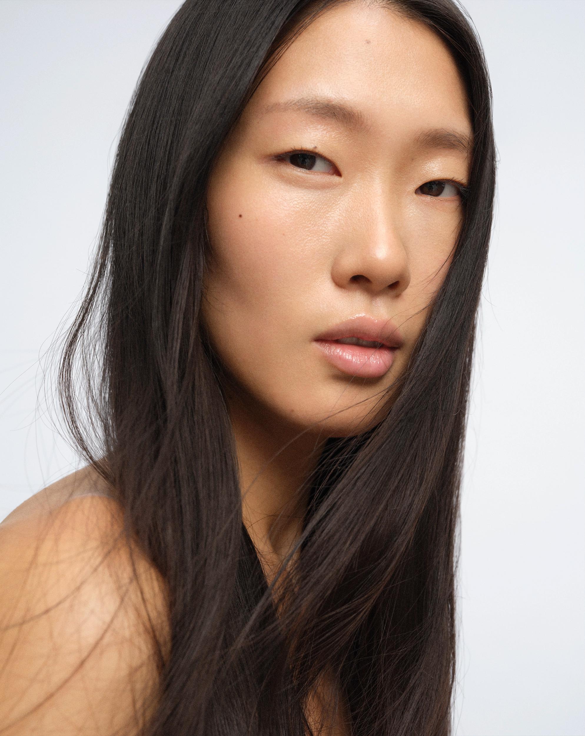 Portrait of a woman with long, straight dark hair falling across her shoulder, dewy glowing skin, and glossy lips, looking at the camera over her bare shoulder