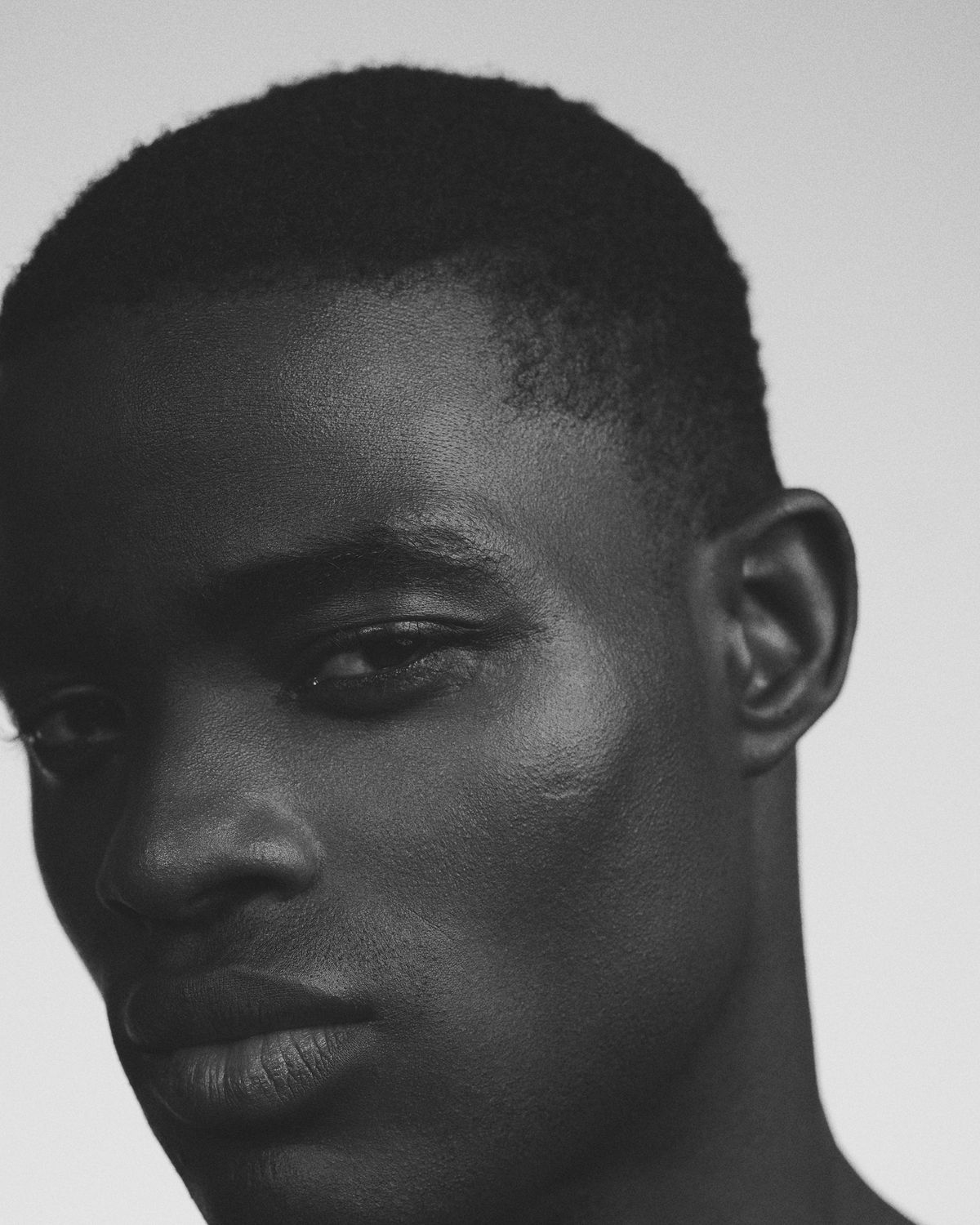 Black-and-white close-up portrait of a young man with short cropped hair and glowing skin, his head tilted down as he looks toward the camera