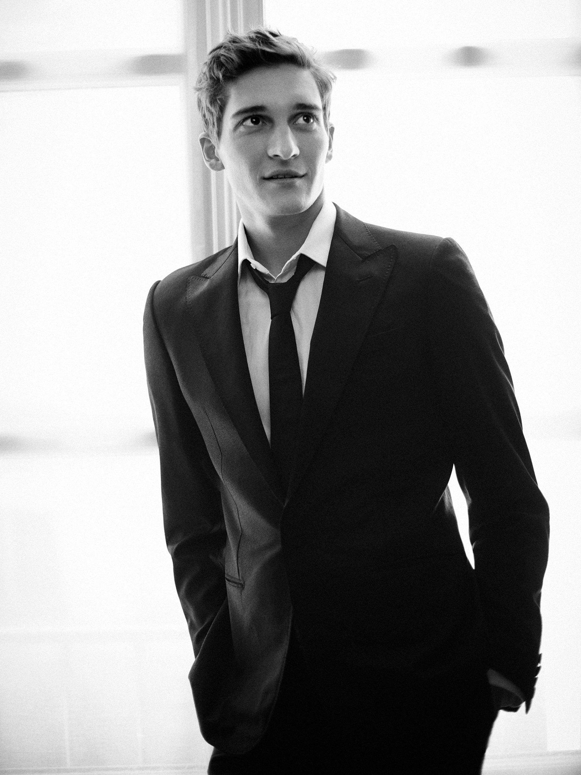 Black-and-white portrait of actor Matvey Lykov in a dark suit and loosened tie, hands in his pockets, glancing up with a slight smile by a bright window