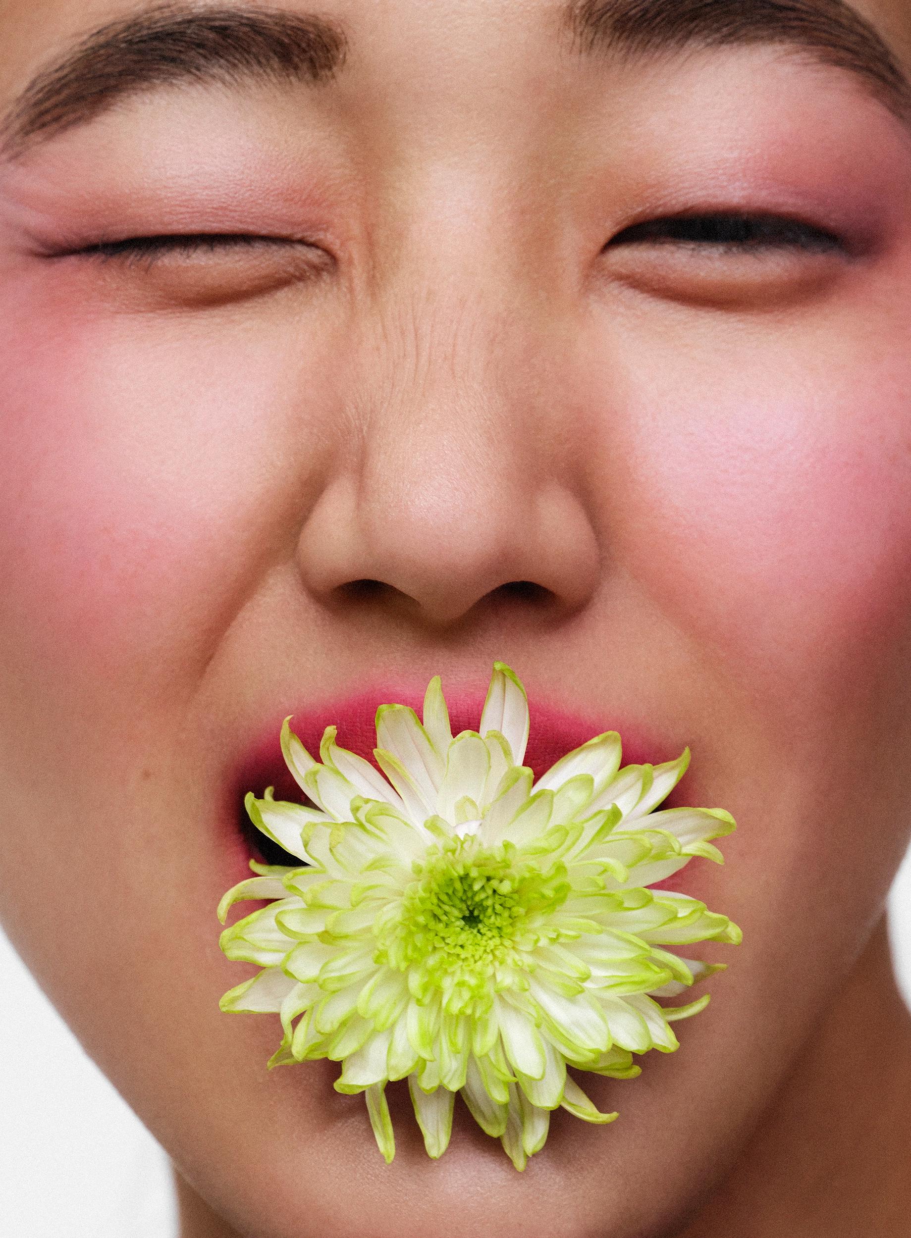 Close-up of a face with eyes squeezed shut, bold pink blush and eyeshadow, and a green-white chrysanthemum held in the mouth.